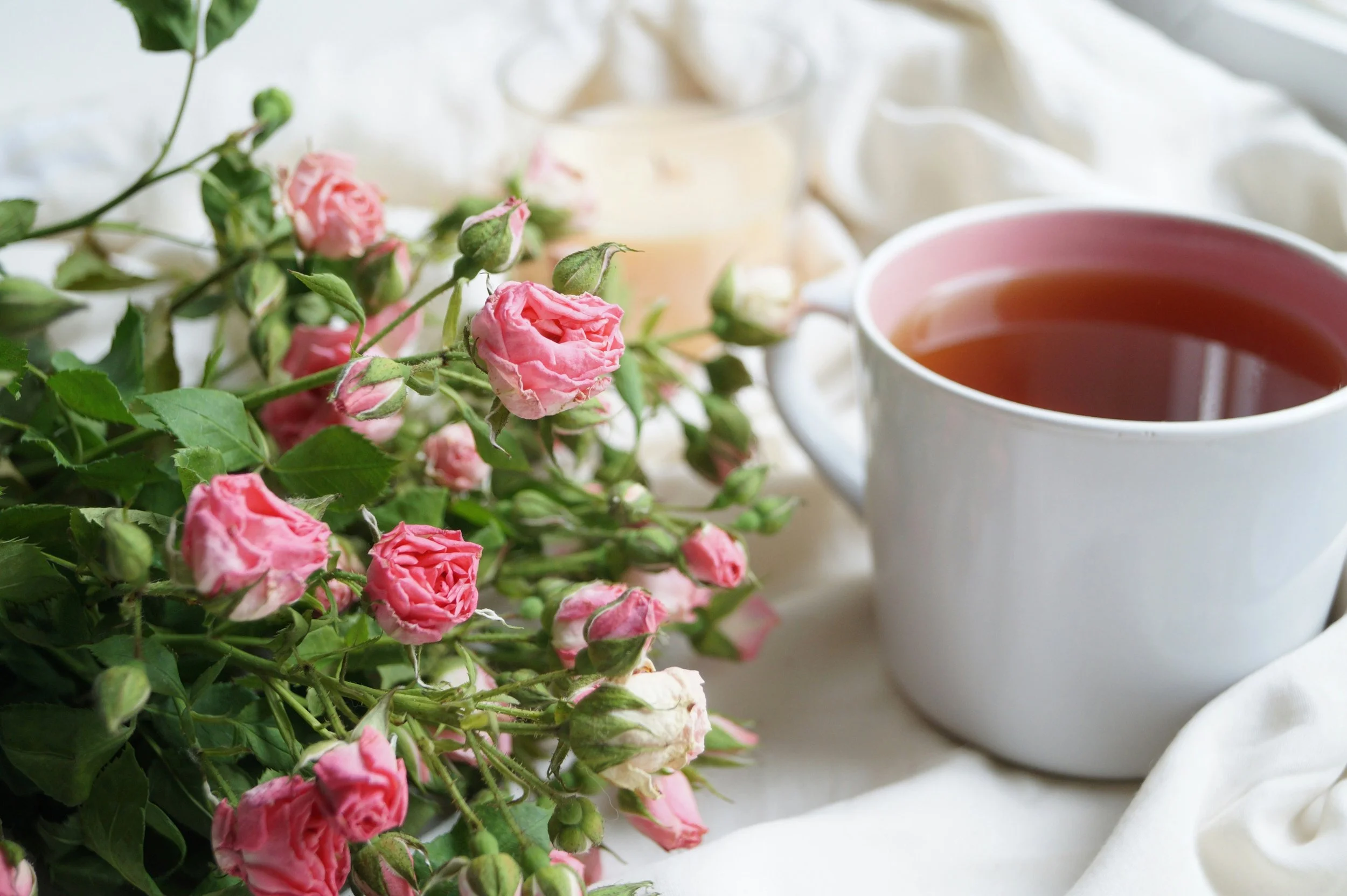 A white cup of tea and pink roses with green leaves on a white table with white fabric in the background.
