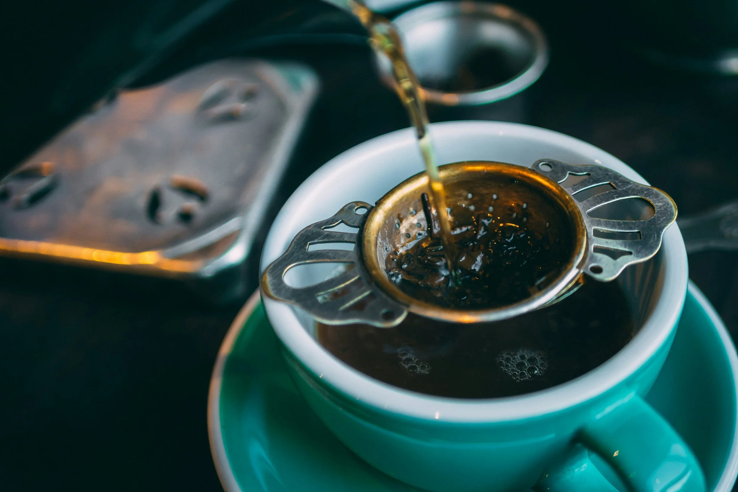 Pouring hot tea through a tea leaf strainer into a teal teacup on a matching teal saucer, with a tea leaf strainer and other tea-steeping accessories in the background.