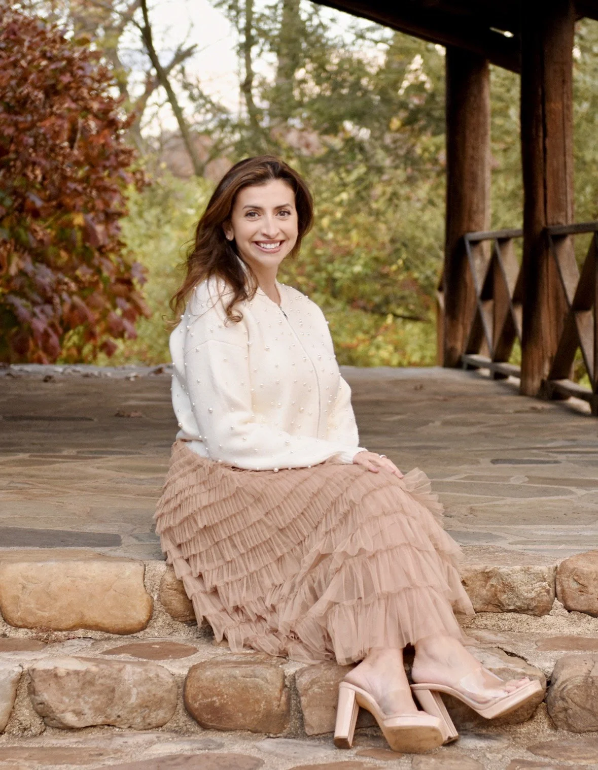 A smiling woman with beautiful brown hair, dressed in a cream-colored blouse and ruffled skirt, sitting on stone steps on a lovely fall day.