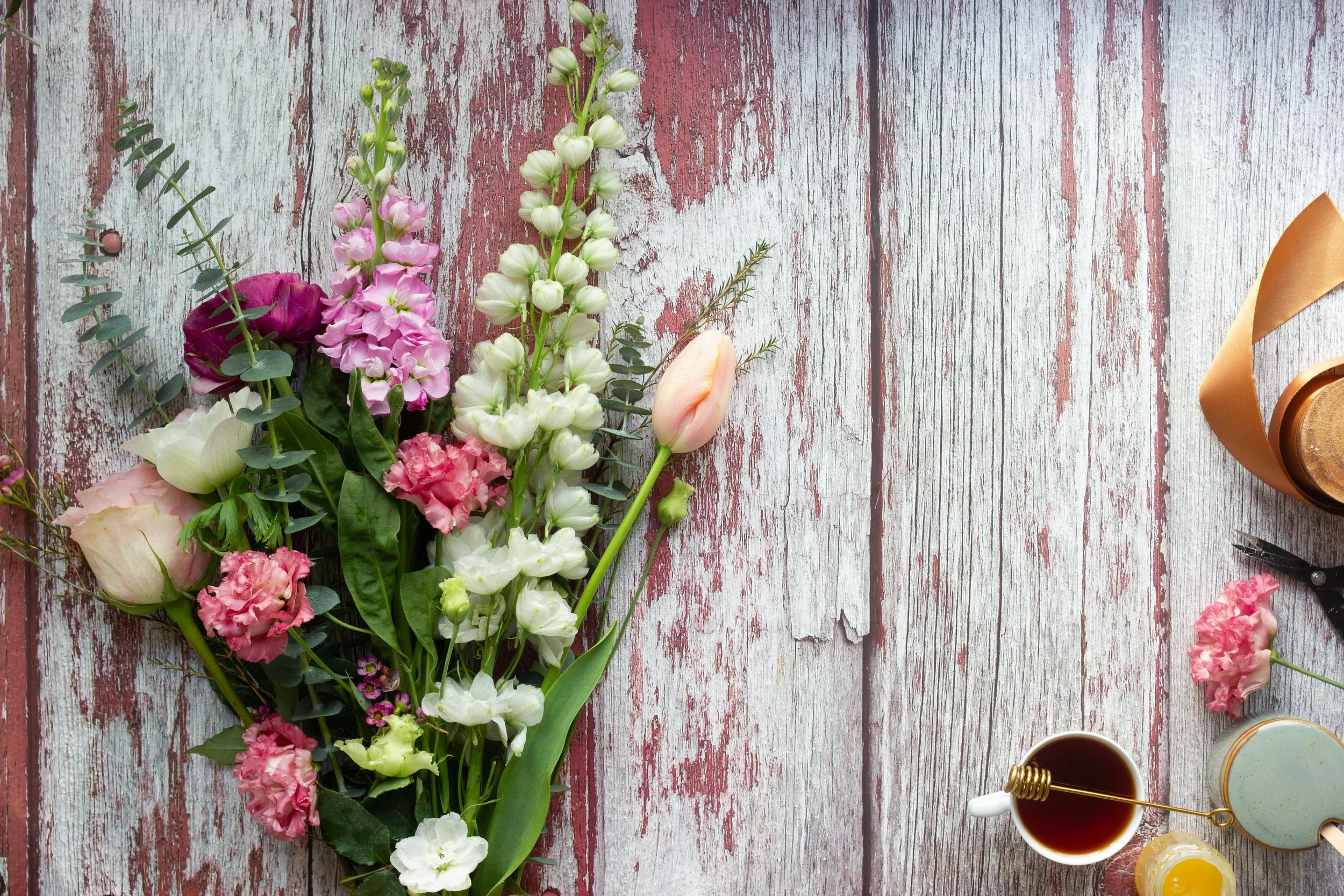 Flat lay of a bouquet of pink and white flowers with green foliage on a distressed white and red wooden surface, with a coffee cup, a pink carnation, a small jar of honey, a cup of tea, a notebook, a pen, and a ribbon on the right side.