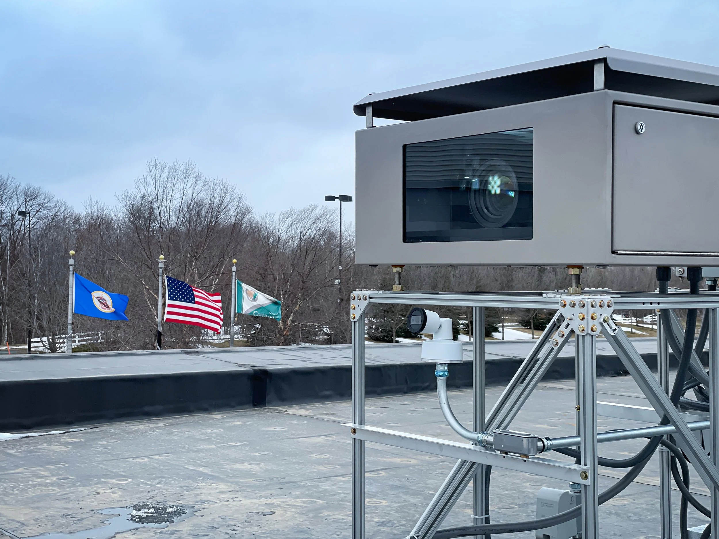 Outdoor projection mapping system in a weatherized enclosure on a rooftop rig, 3 flags behind it under gray sky