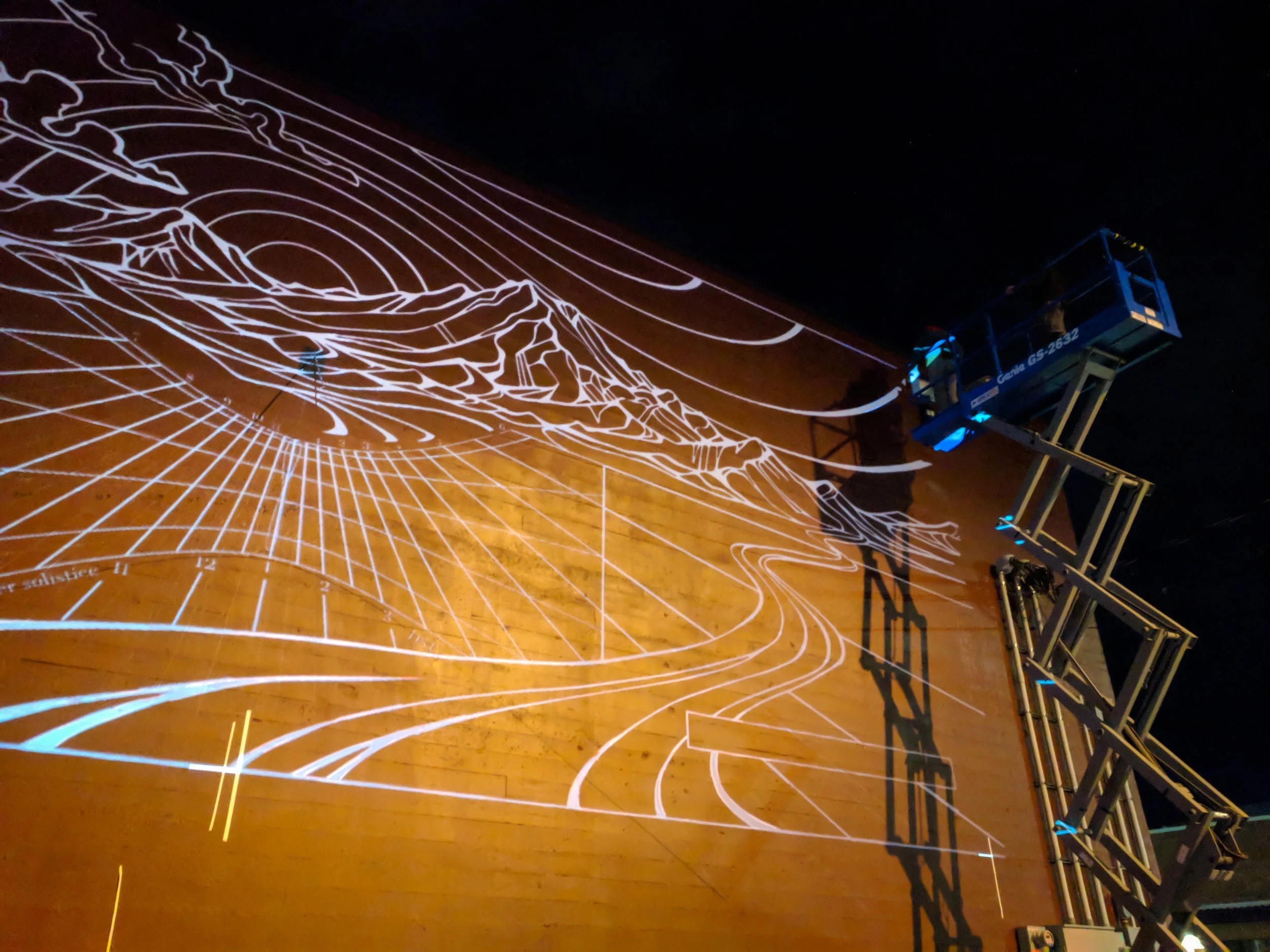 Close angle of sundial hour lines and mountain linework projected on a wall, tracing crew working from a boom lift
