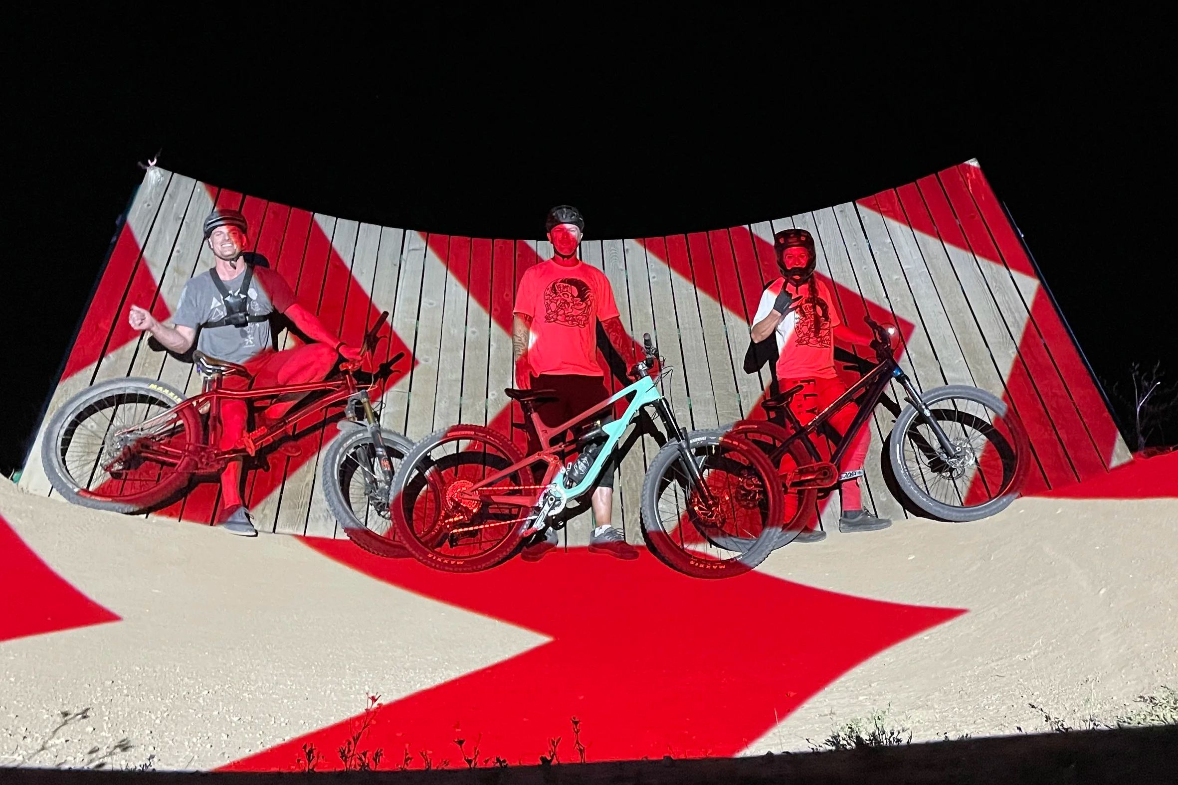 3 riders line up with bikes below a chevron mapped wall ride in Boise, red projection washing over the group.