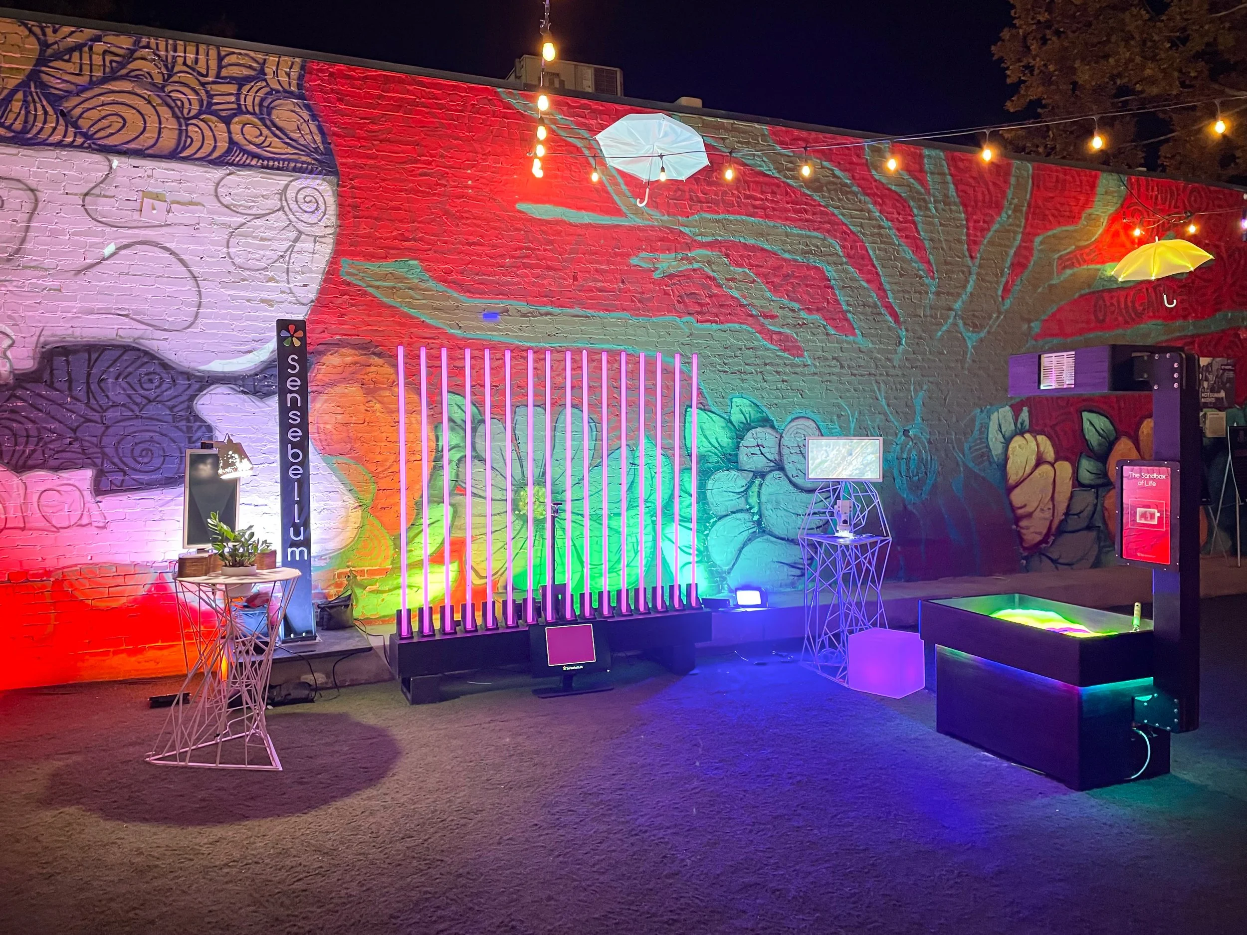 Interactive light installations line the mural wall at 5000 Watts at night, LED bars glowing pink over green turf