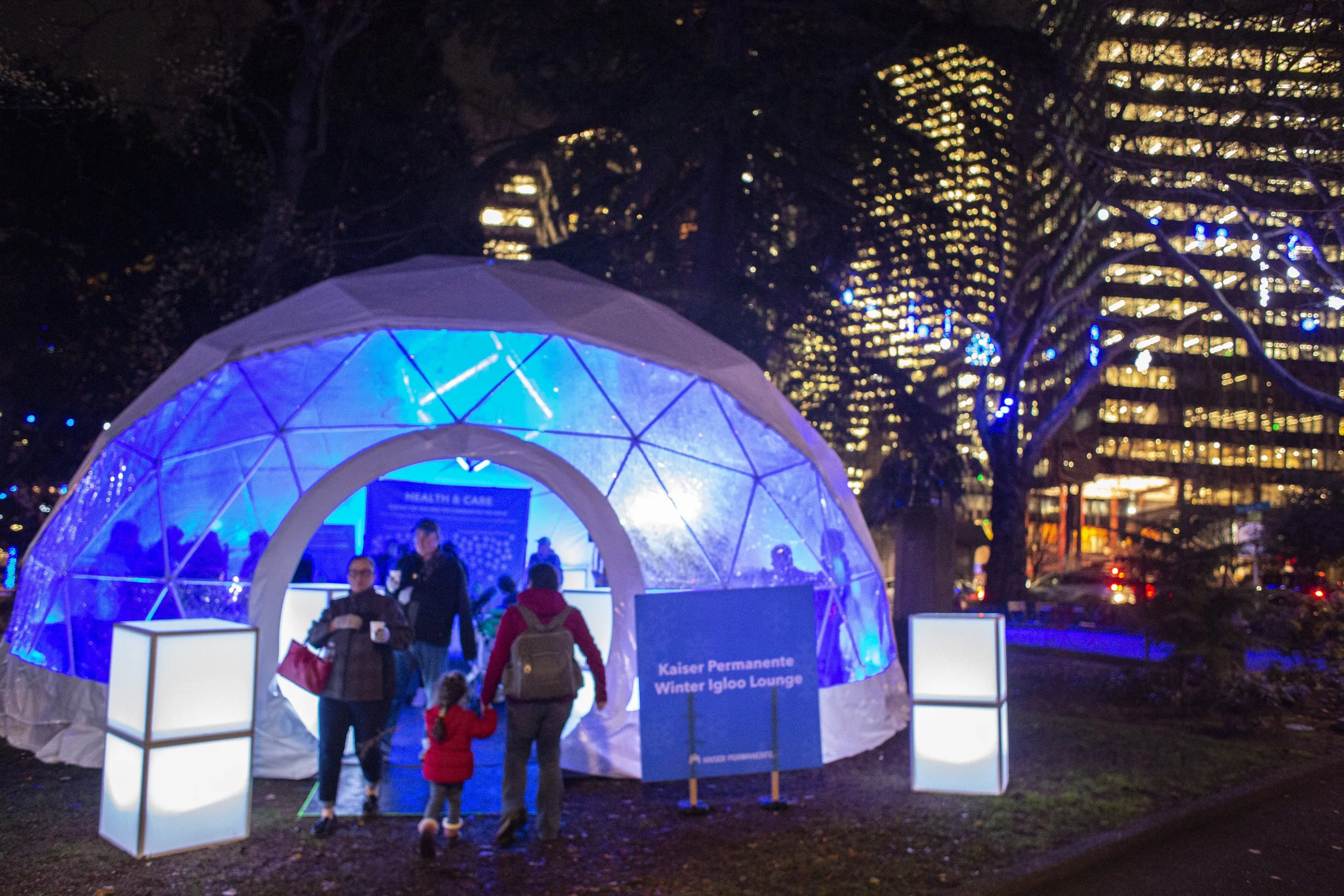 Geodesic event dome glowing blue at night in Denny Park, Seattle, with families entering past 2 luminous cube pedestals