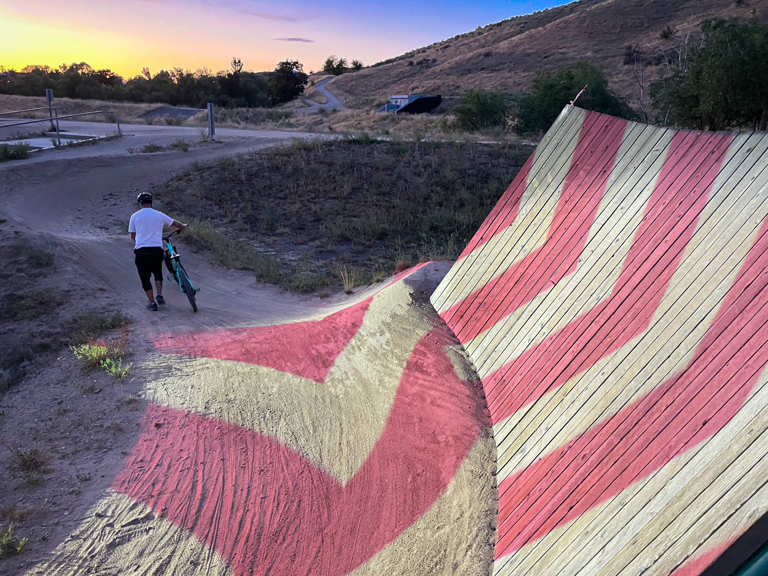 Rider walks a bike past the chevron mapped wall ride at sunset, projected stripes faint in the last daylight.