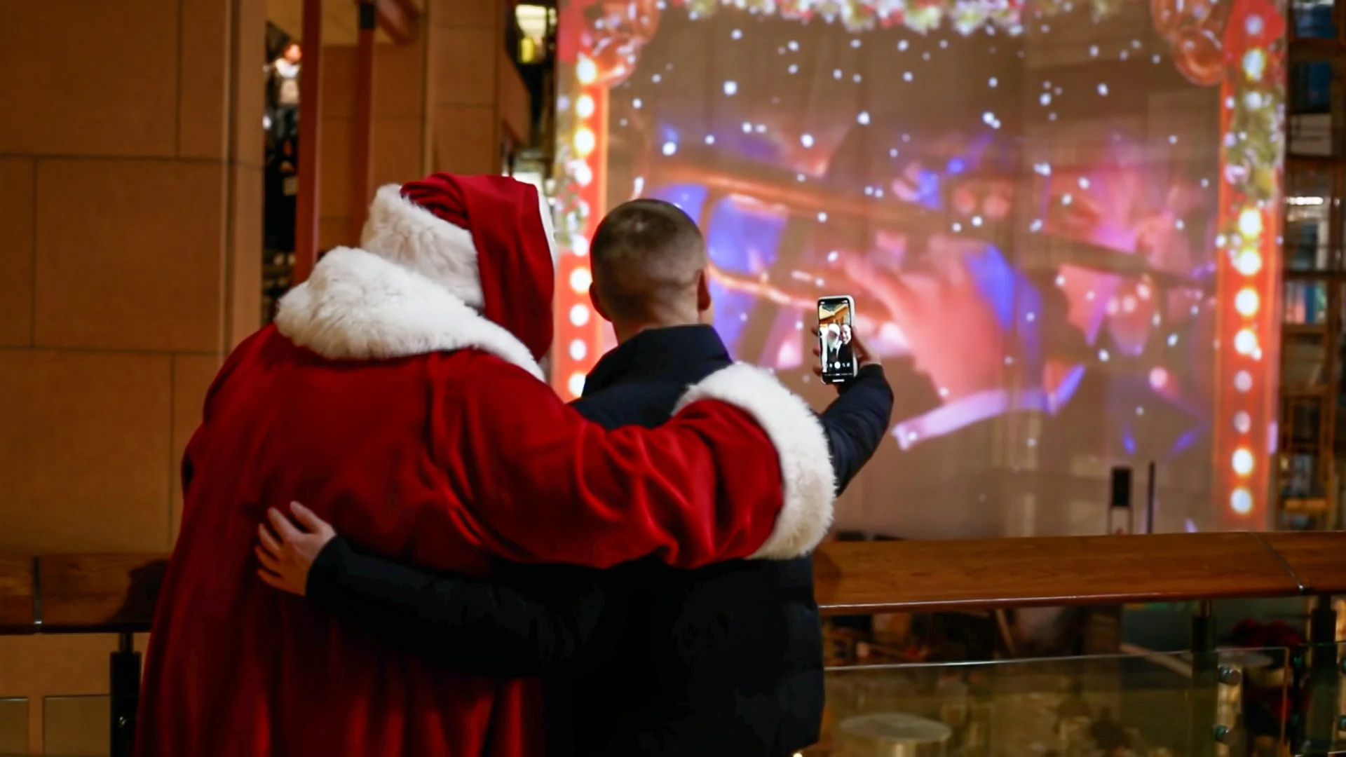 Santa and a guest taking a selfie in front of the sing along projection at Pacific Place, Seattle, snow falling on screen.