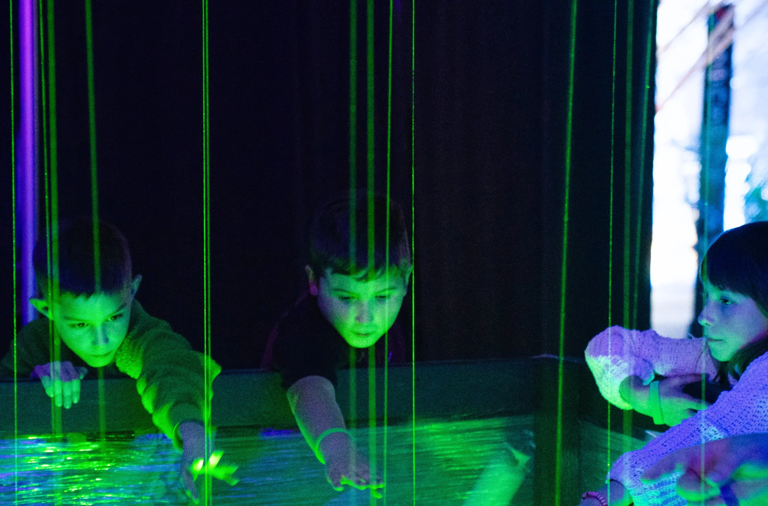 3 children reach into green laser harp beams at a NOVA Light Labs installation, faces lit by the glow