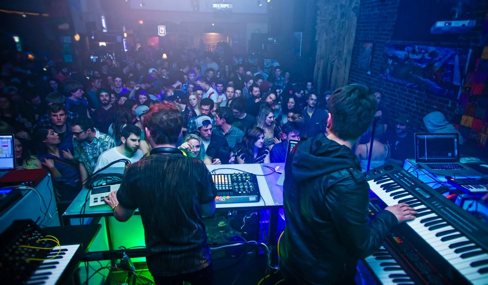 Live From Space performers at keyboards and drum machines facing a packed crowd under blue and green light