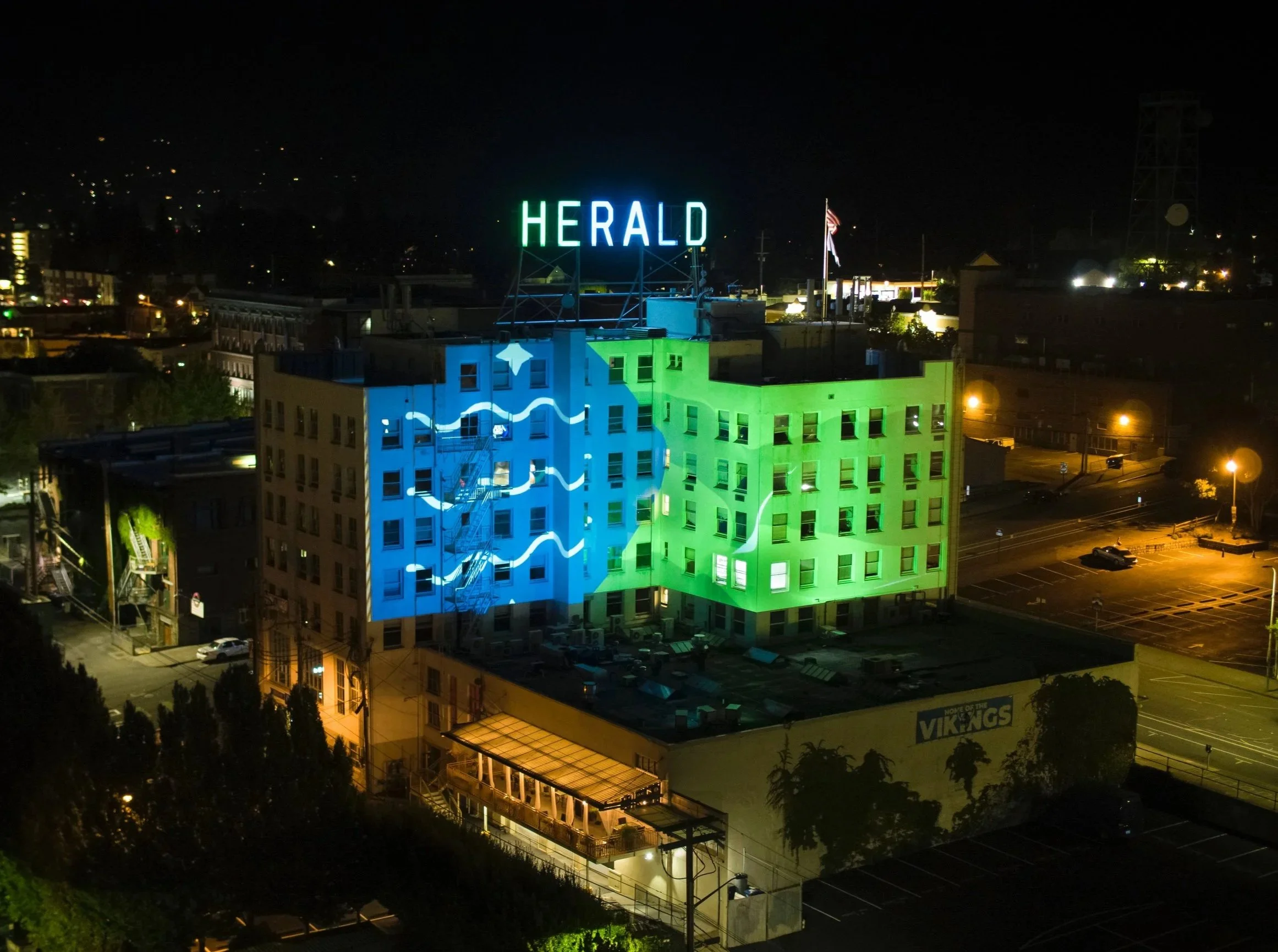 The Herald projection mapping in Bellingham, facade split blue and green with flowing white lines at night.