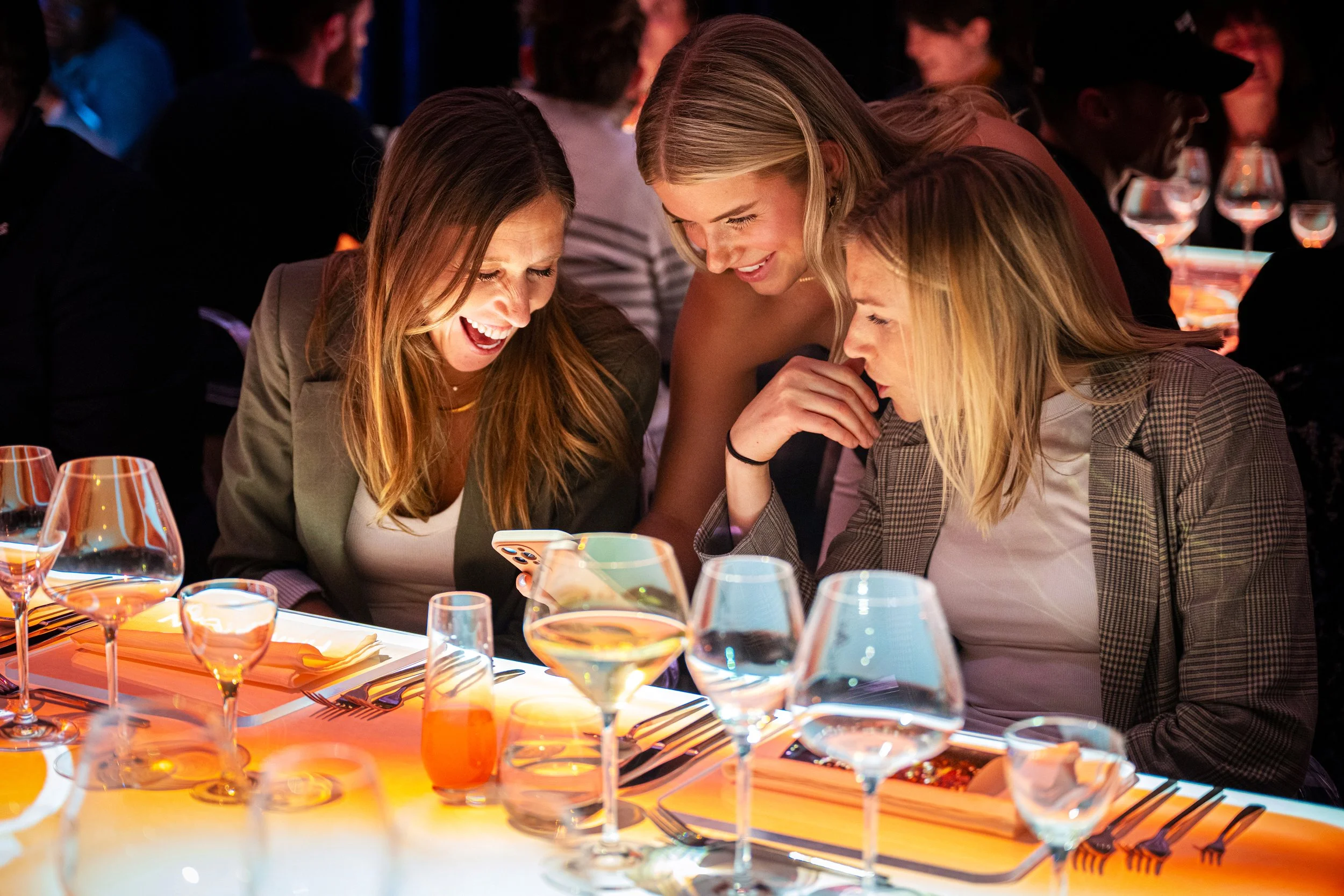 The Radiant Table immersive dining, three guests lean in over a phone at the amber projection mapped table