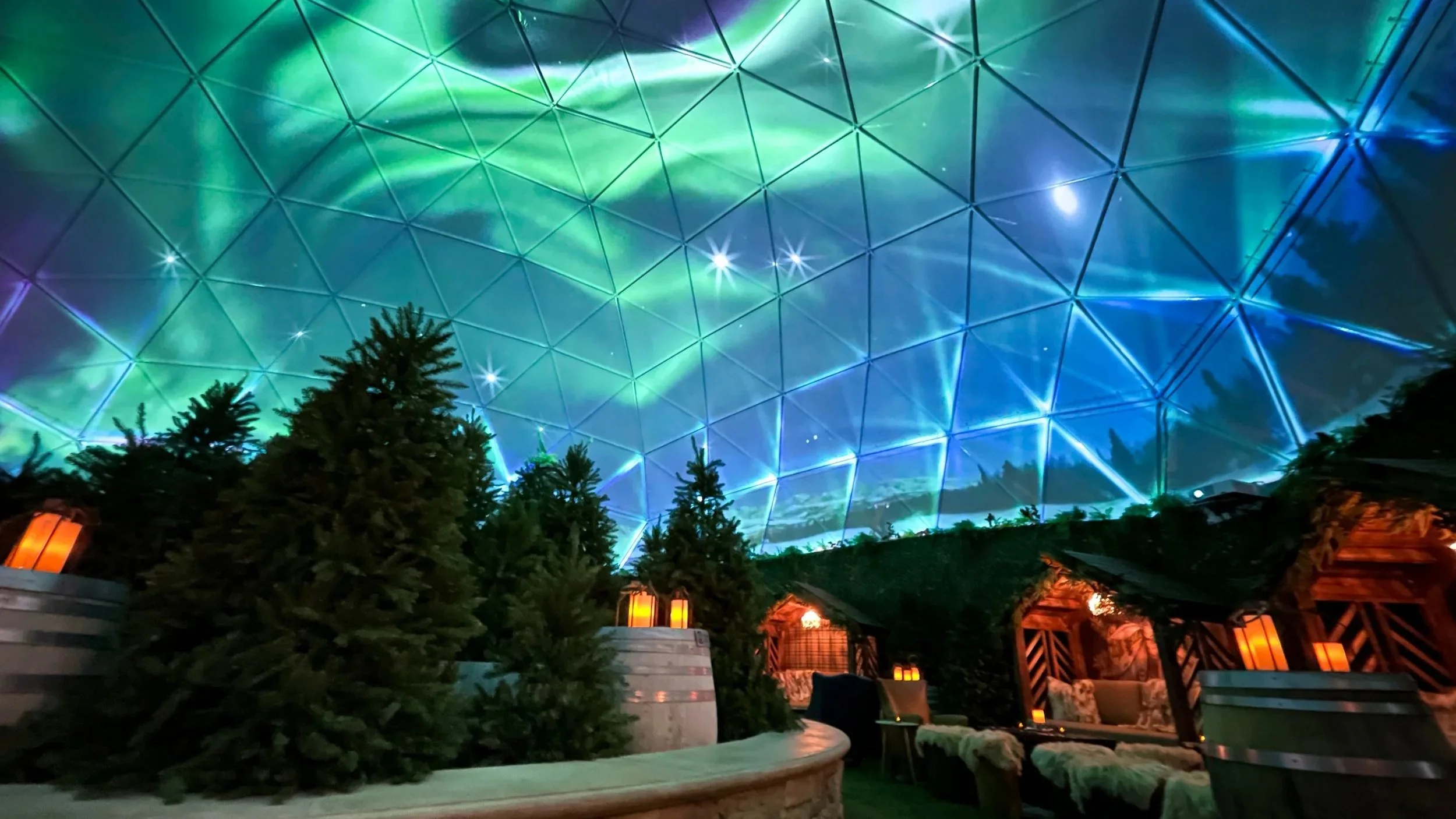 Projection mapping in a dome, green aurora and stars across a geodesic ceiling above pine trees and lantern lit cabins.