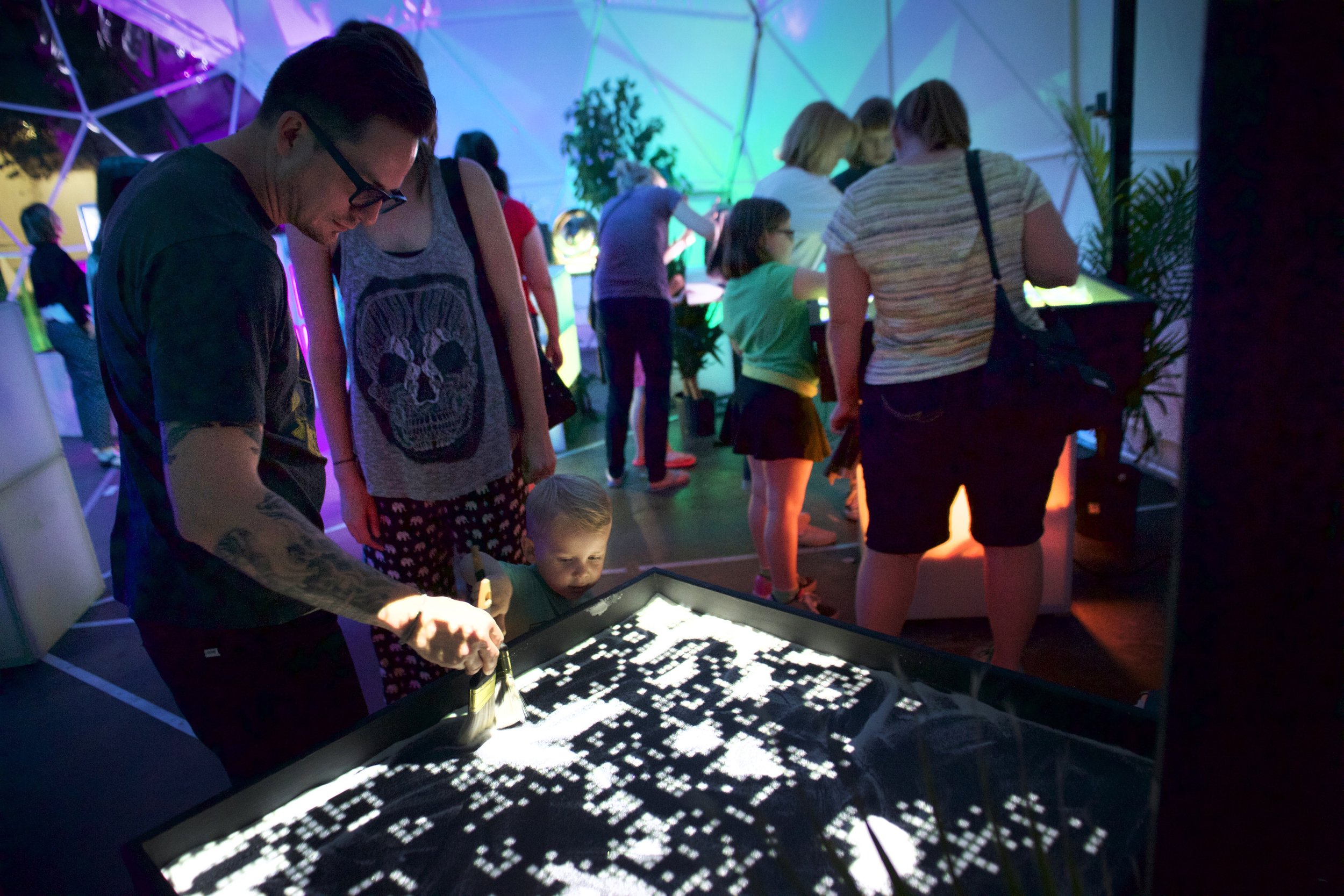 Guests brushing sand on The Sandbox of Life inside The Sensatorium dome in Mesa, Arizona, projected terrain glowing white.