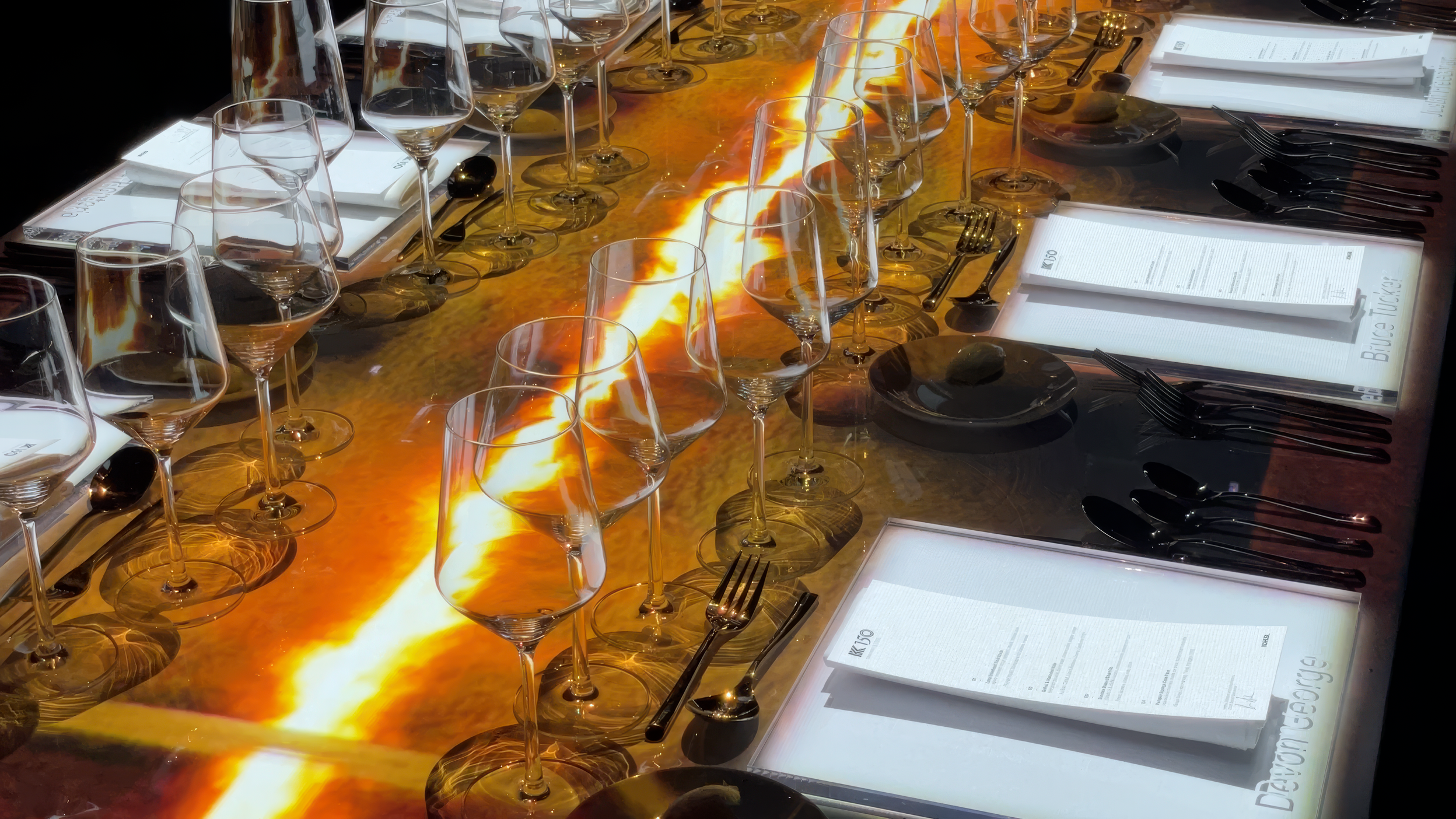 Immersive projection dining, a molten amber river of light down a set table between wine glasses and menus.