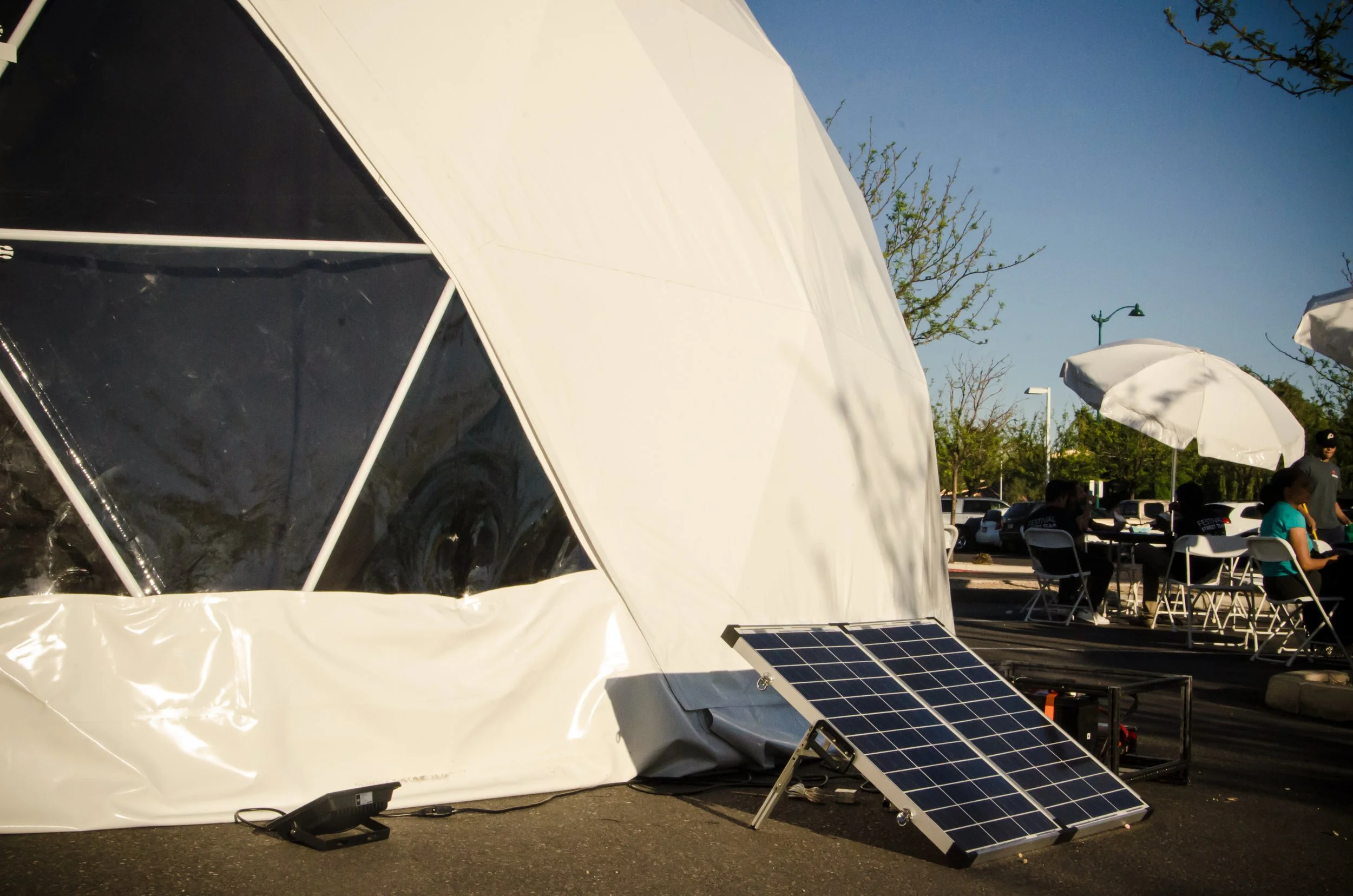 The Sensatorium dome exterior by day in Mesa, Arizona, a folding solar panel propped at its base beside festival tables.