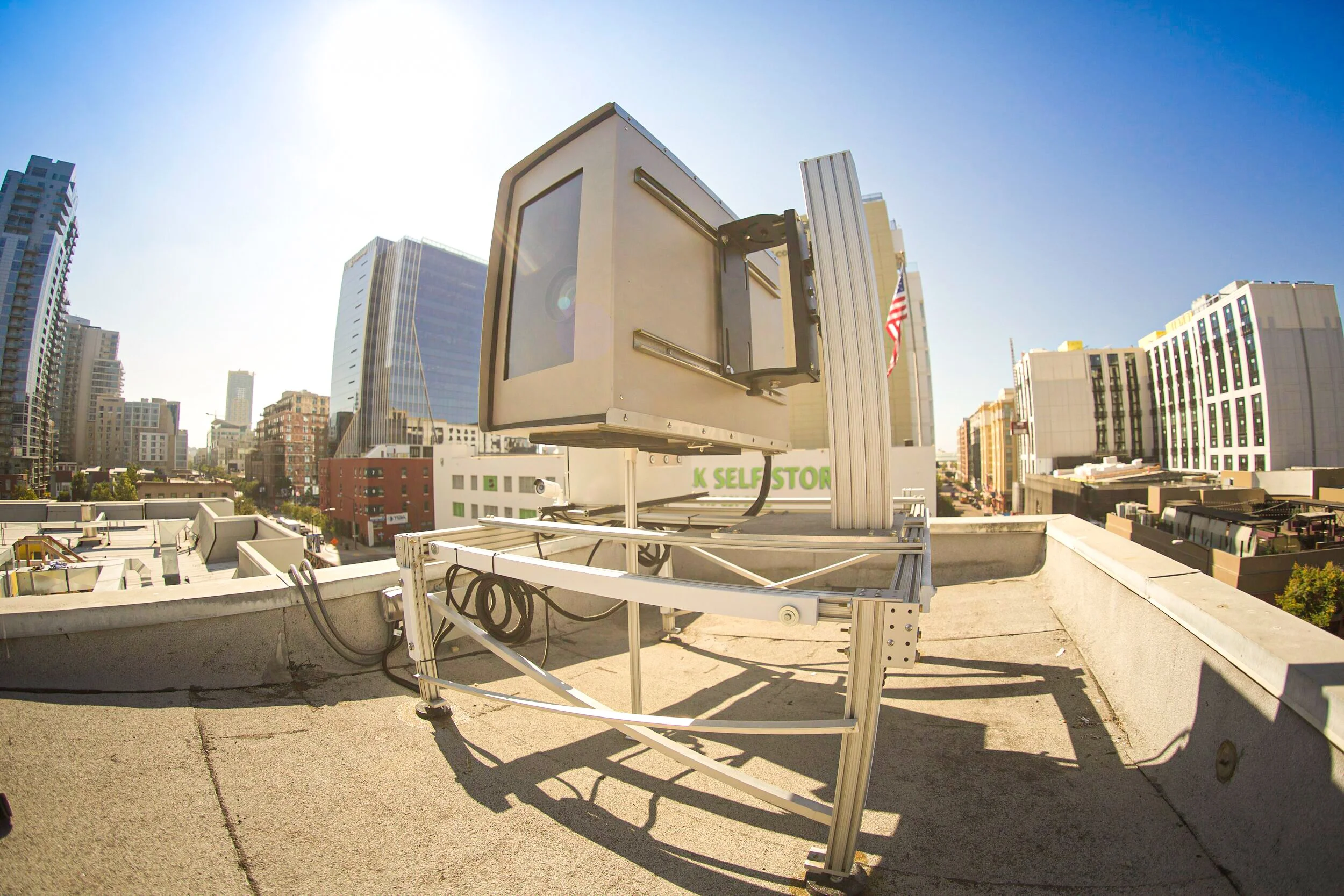 Permanent projection mapping system mounted on an open metal rig atop a city rooftop, lens angled over the skyline