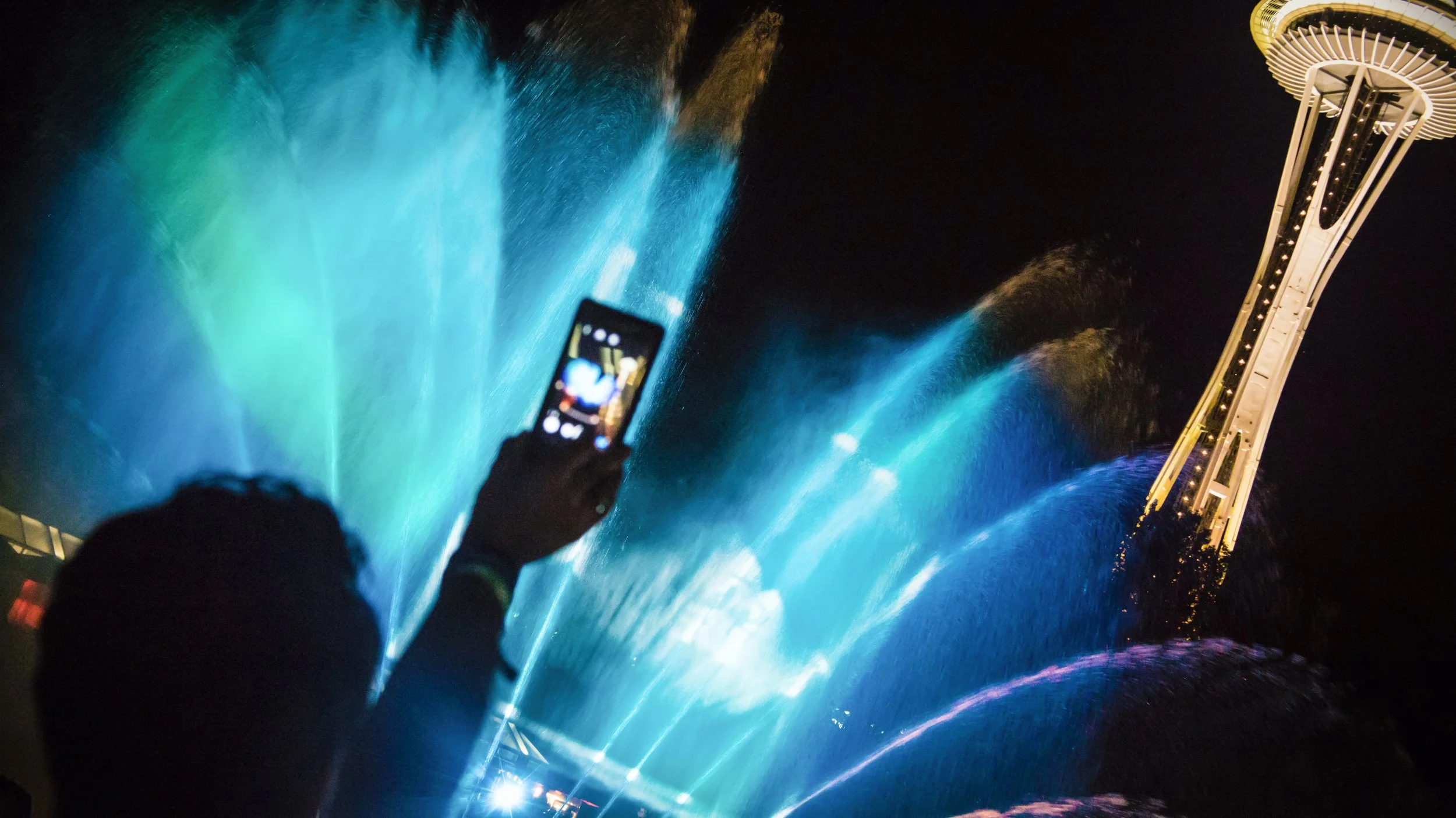Bumbershoot International Fountain projection mapping, guest films blue lit water with phone below Space Needle