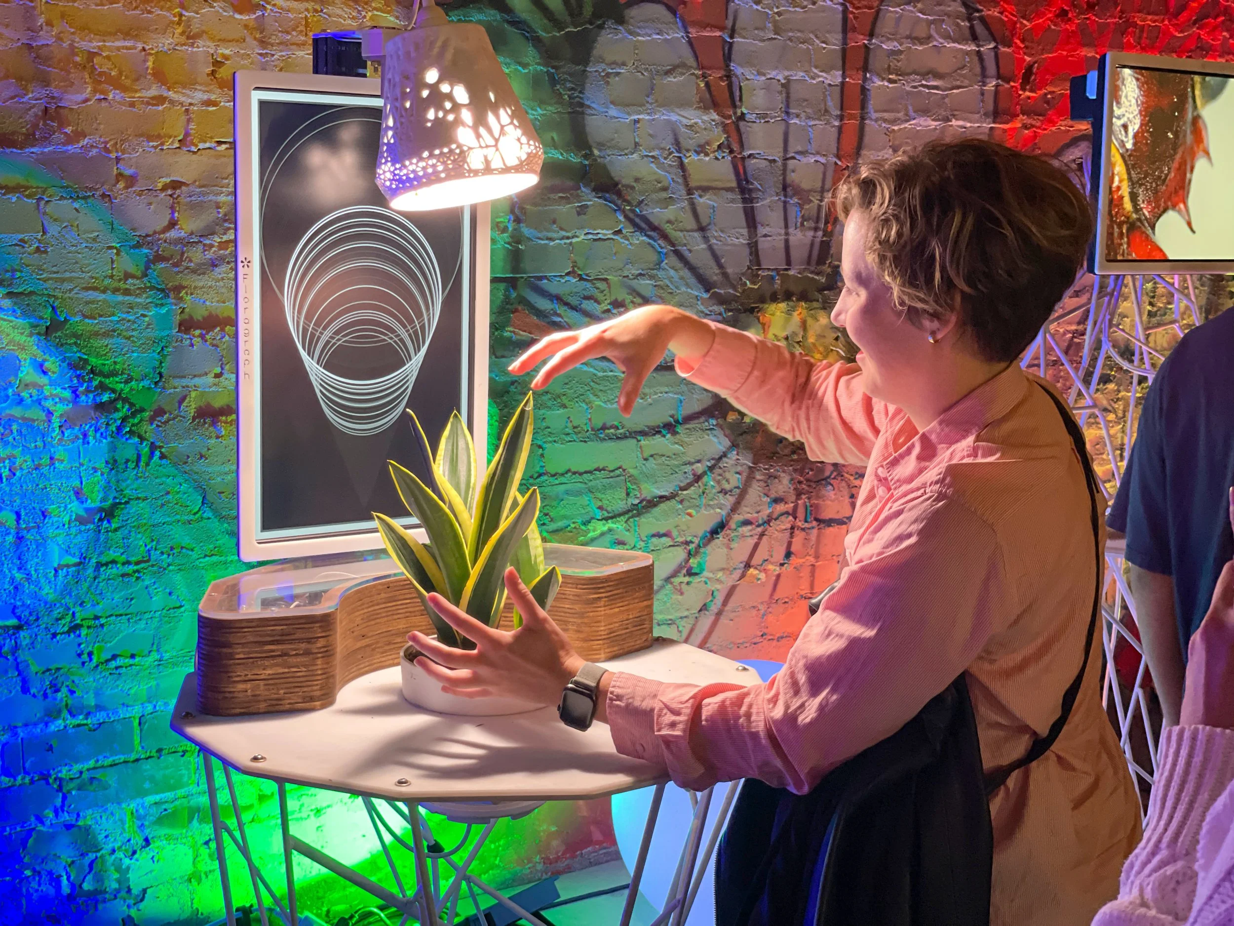 Guest reaches over a live plant at the Floragraph installation at 5000 Watts, brick wall lit green and blue behind her