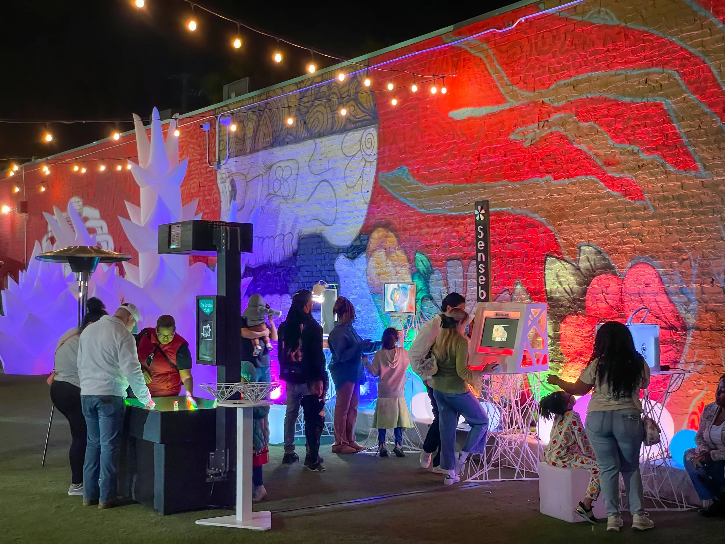 Guests play interactive art installations at 5000 Watts at Esther's Park in Sacramento under a mural lit in red and blue