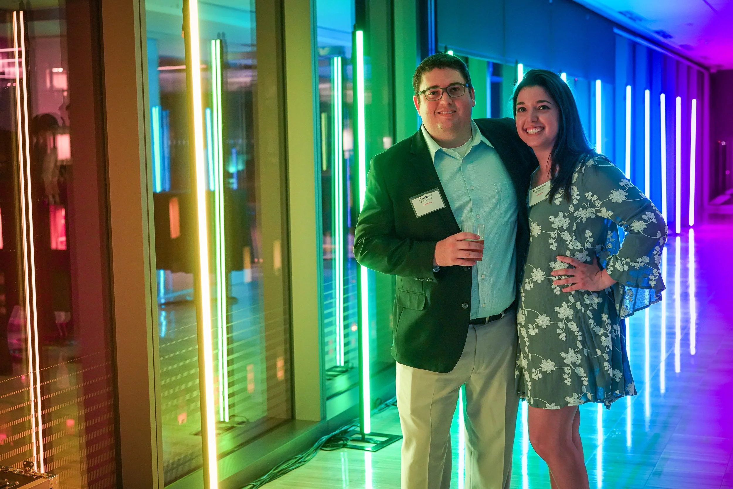 2 guests pose before rainbow LED bars at Holland & Knight in Washington, DC, green and blue light across the glass.