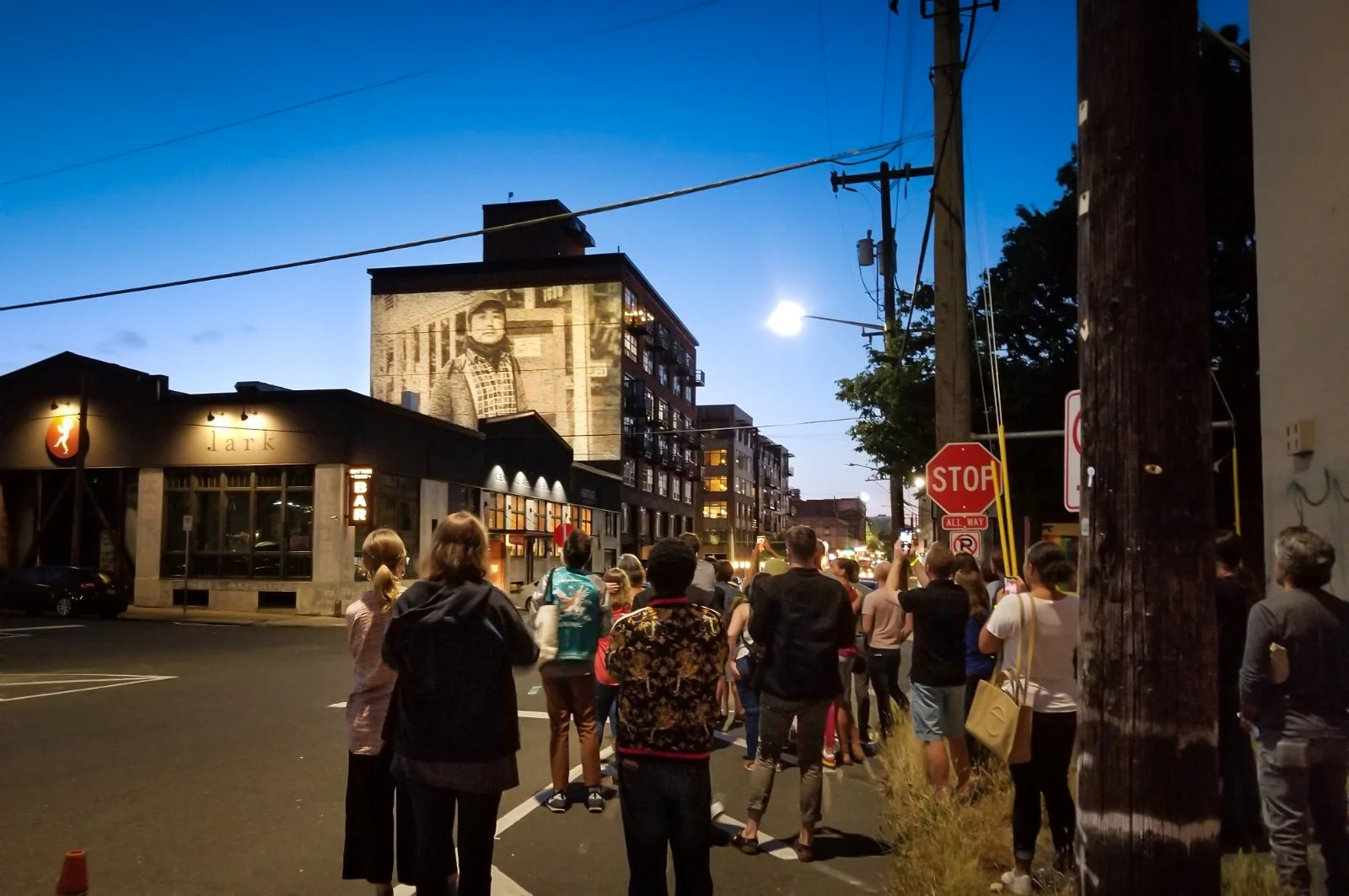 Woodworth Apartments projection mapping, Capitol Hill Seattle, crowd gathered in the street watching a portrait at dusk.
