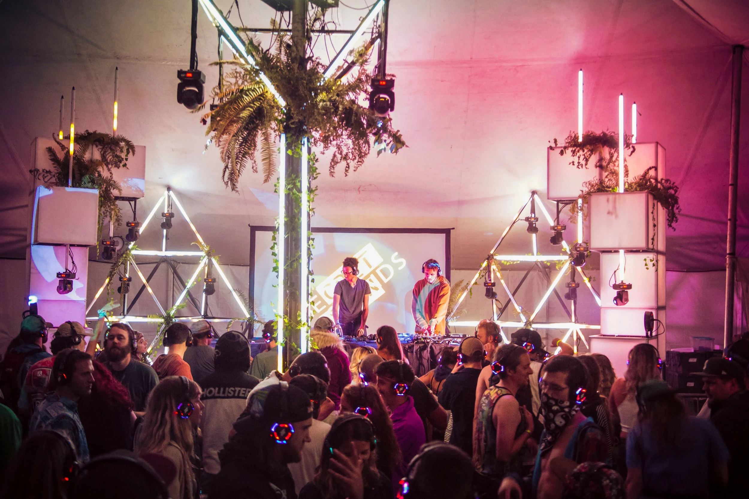 Lighting design for a silent disco stage, illuminated triangle trusses framing DJs over a headphone crowd
