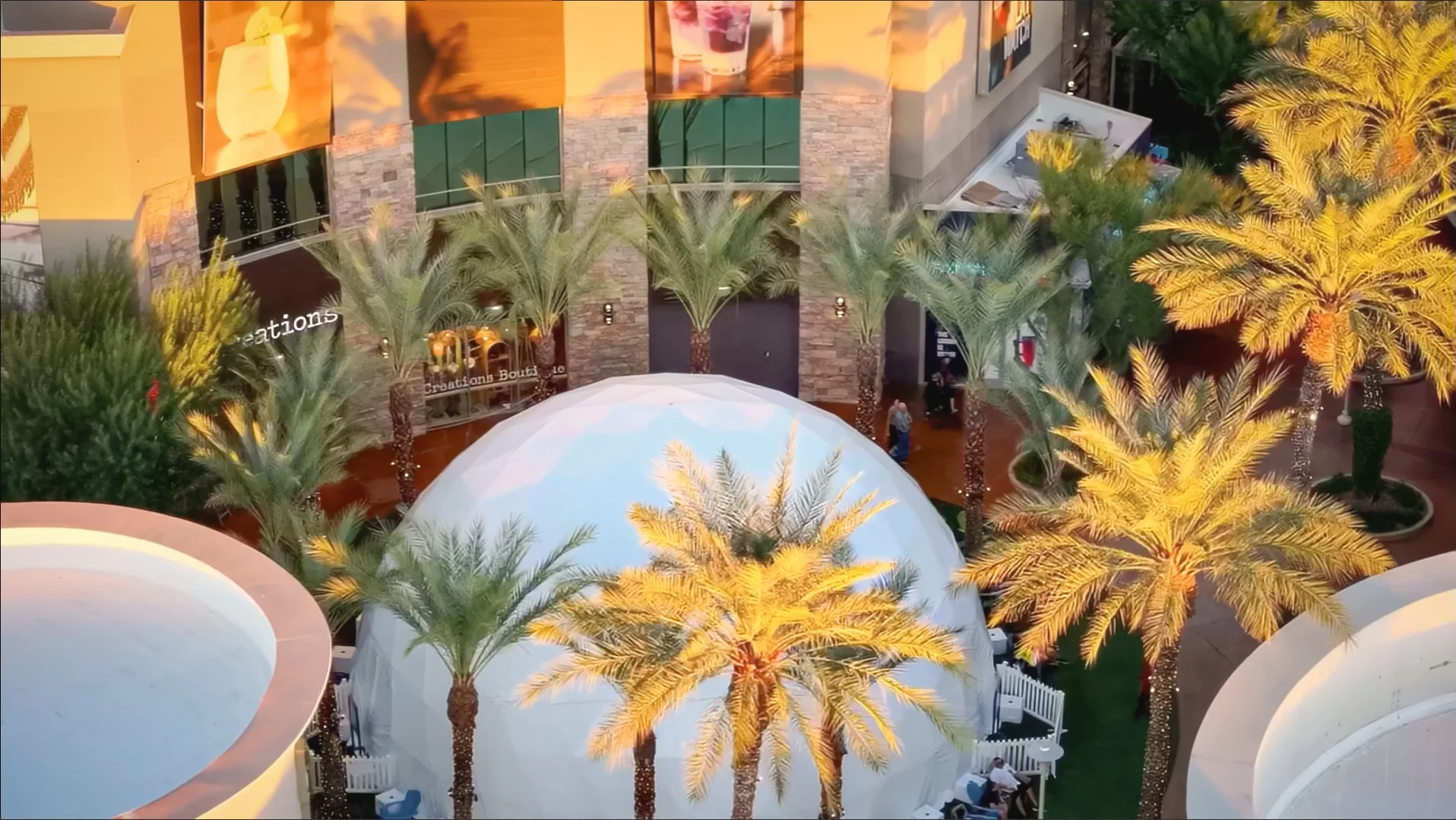 Aerial view of a white geodesic event dome set among palms at an outdoor retail plaza before showtime.