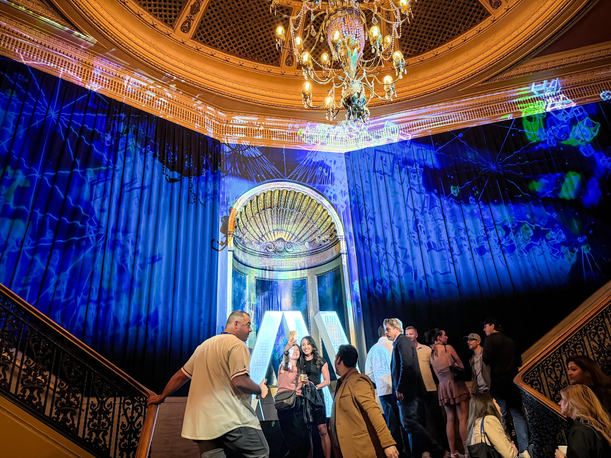 Guests crowd the Warfield balcony landing in San Francisco as blue projection mapping covers the drapery and chandelier.