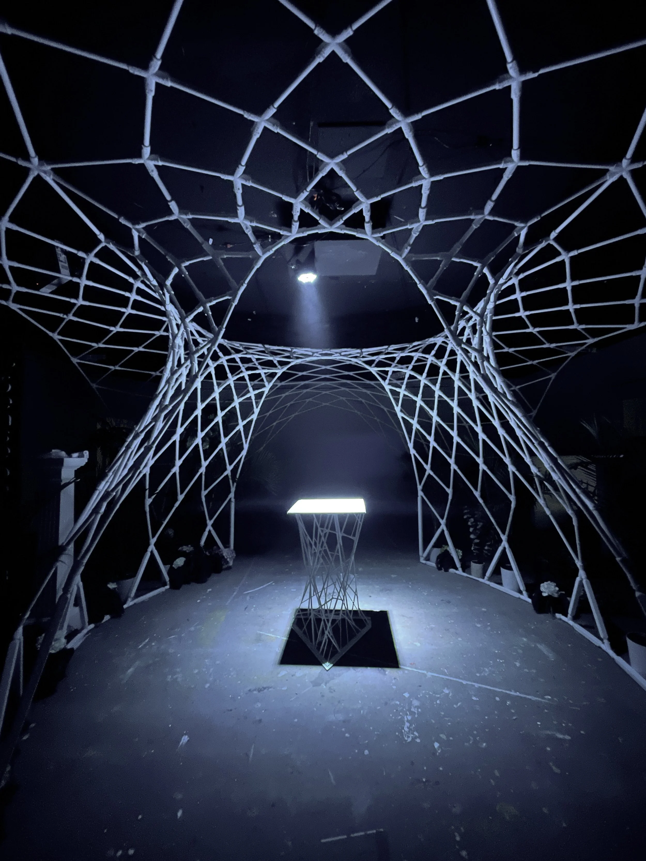 Parametric mesh pavilion lit in the SKIP building at JUMP in Boise, Idaho, a single glowing table at its center.