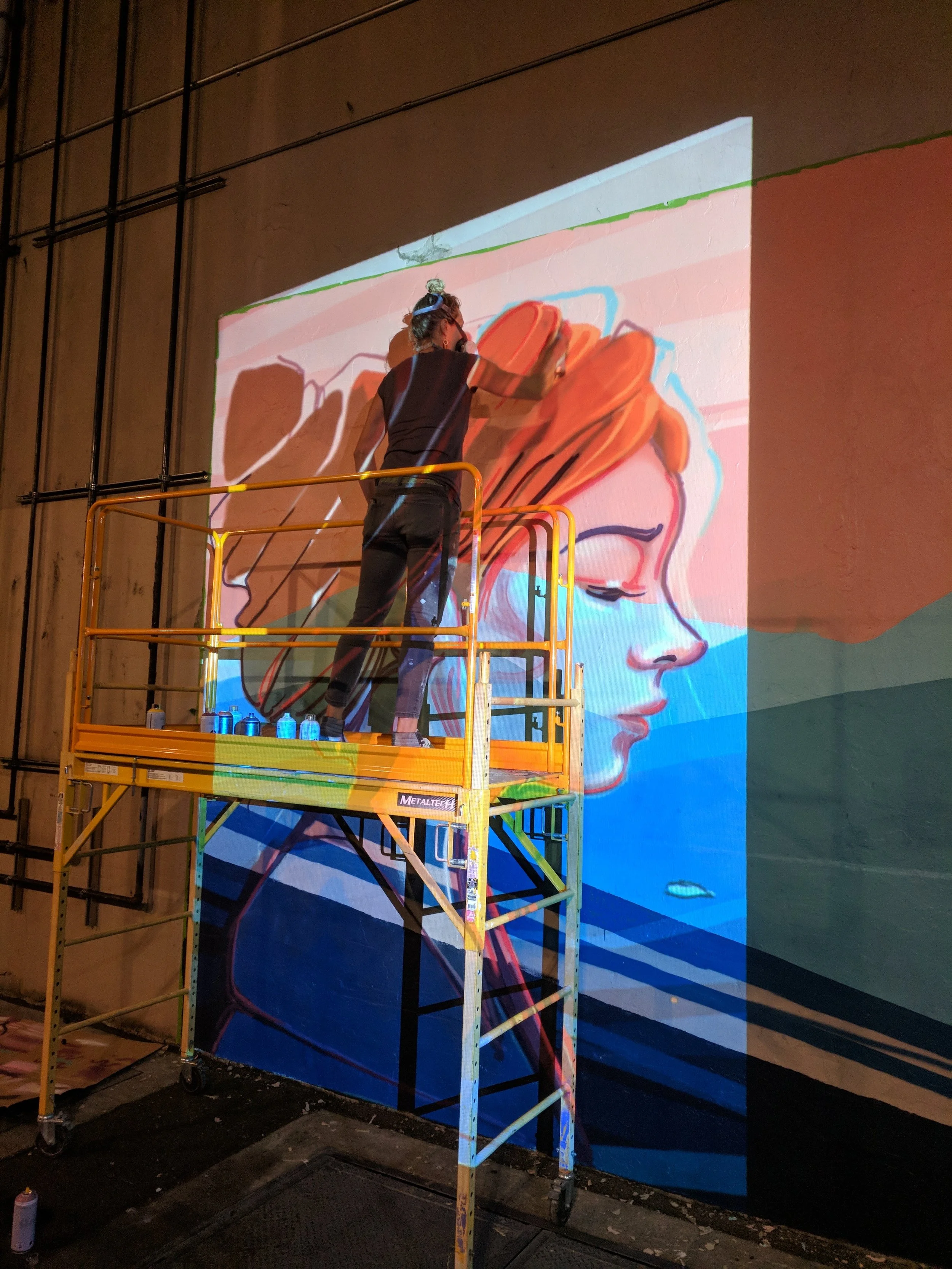 Muralist on a scaffold paints a woman's portrait over a projected design at night, spray cans lined along the rail
