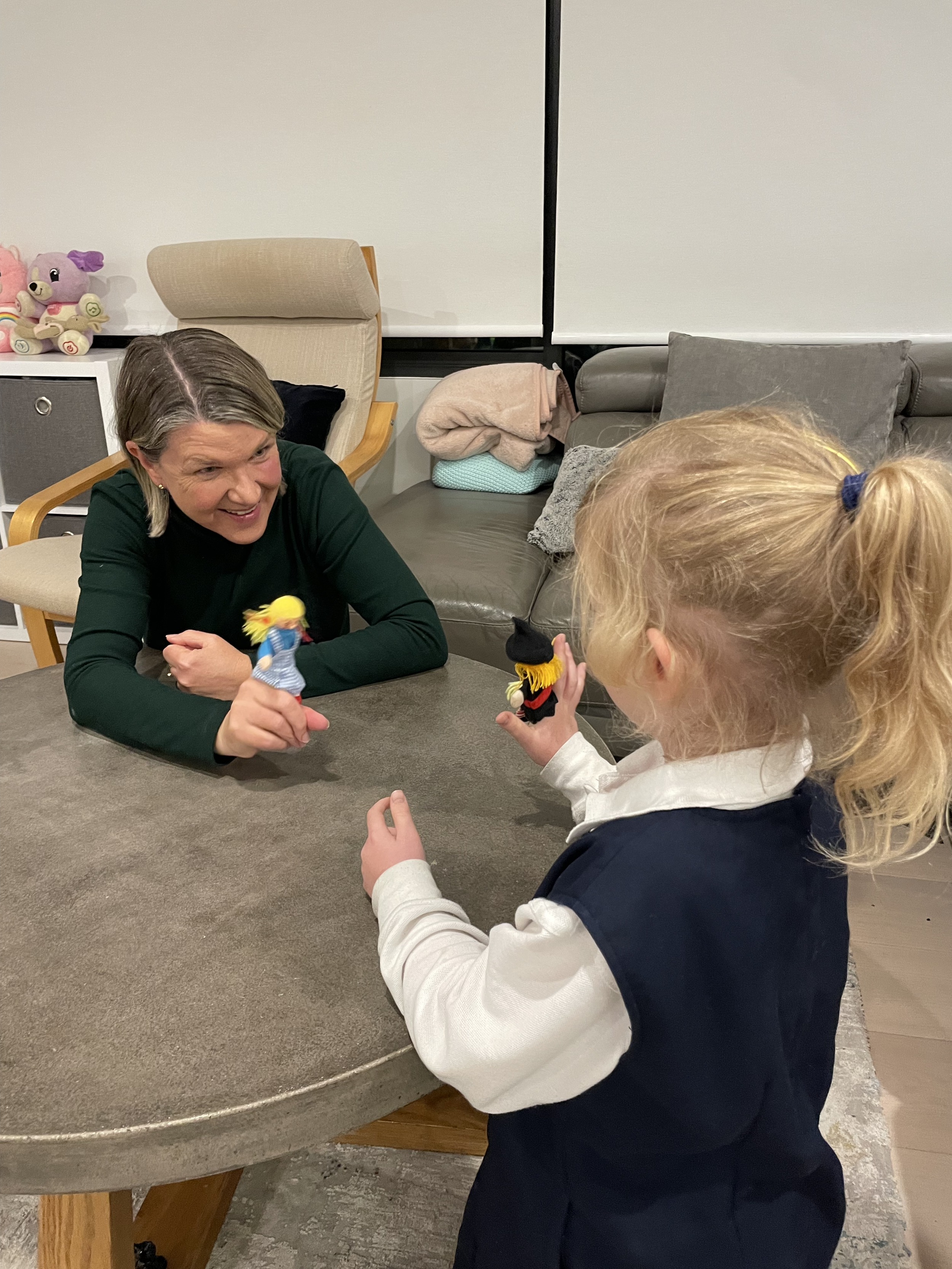 A woman and a young girl are playing with puppets at a table indoors. The woman is smiling and holding a puppet, while the girl is holding a puppet and facing the woman.