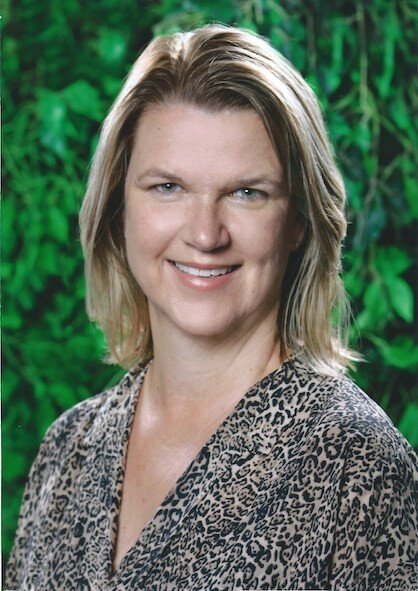 A woman with shoulder-length blonde hair smiling, wearing a leopard-print top, standing in front of a green leafy background.