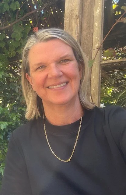 A smiling woman with shoulder-length gray hair wearing a black shirt and a gold necklace, outdoors with greenery and a wooden post in the background.