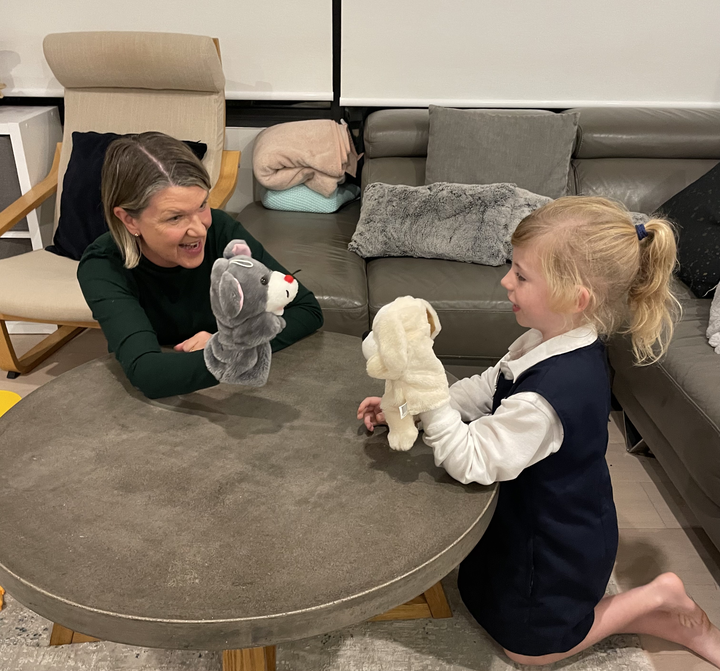 A woman and a young girl sitting at a round table playing with plush puppets, smiling and engaging in a puppet show.