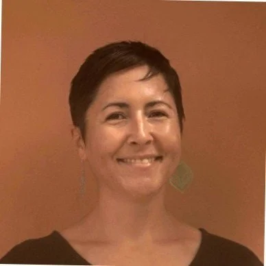 A woman with short dark hair smiling in front of a brown wall.