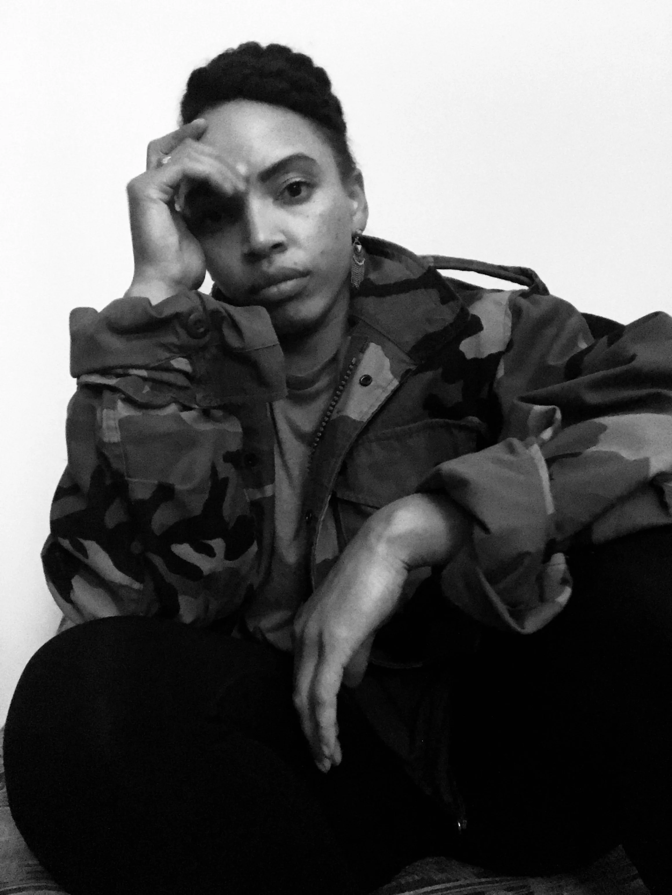 A person with short hair, wearing a camouflage jacket, sitting with one hand resting on their face and the other hand on their knee, looking directly at the camera with a serious expression.