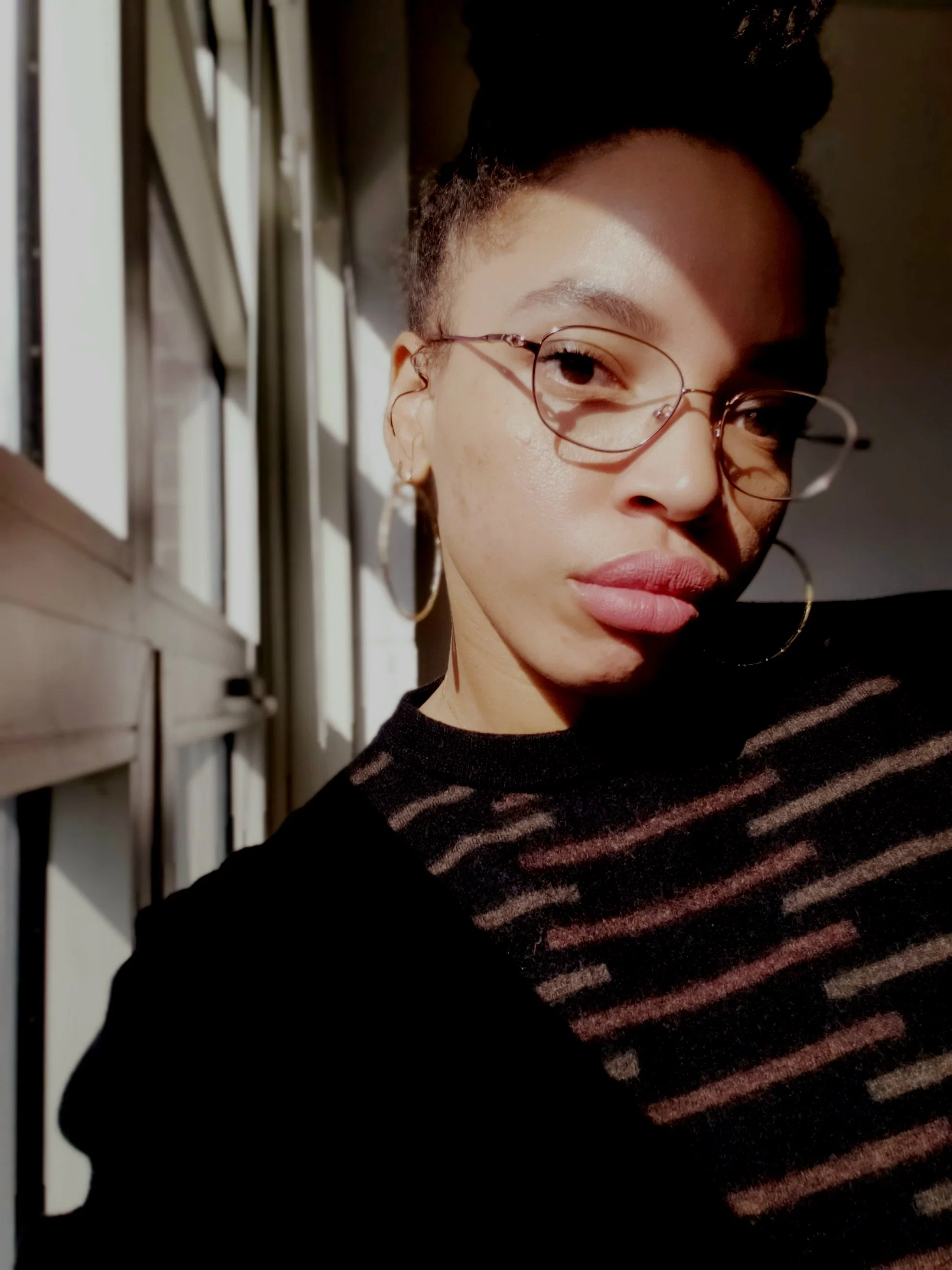 Close-up of a woman with glasses, hoop earrings, and dark wavy hair styled in a bun, standing by a window with sunlight and shadows on her face and shirt.
