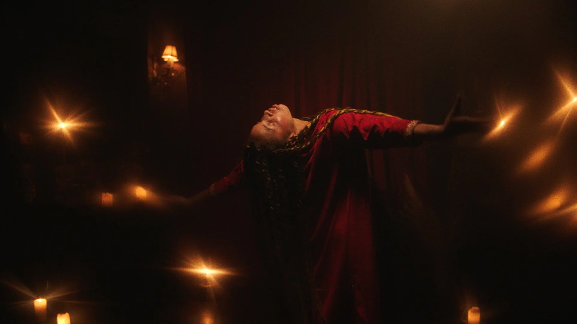A person with braided hair in a red dress standing in a dark room with arms outstretched, illuminated by warm candlelight and wall sconces.
