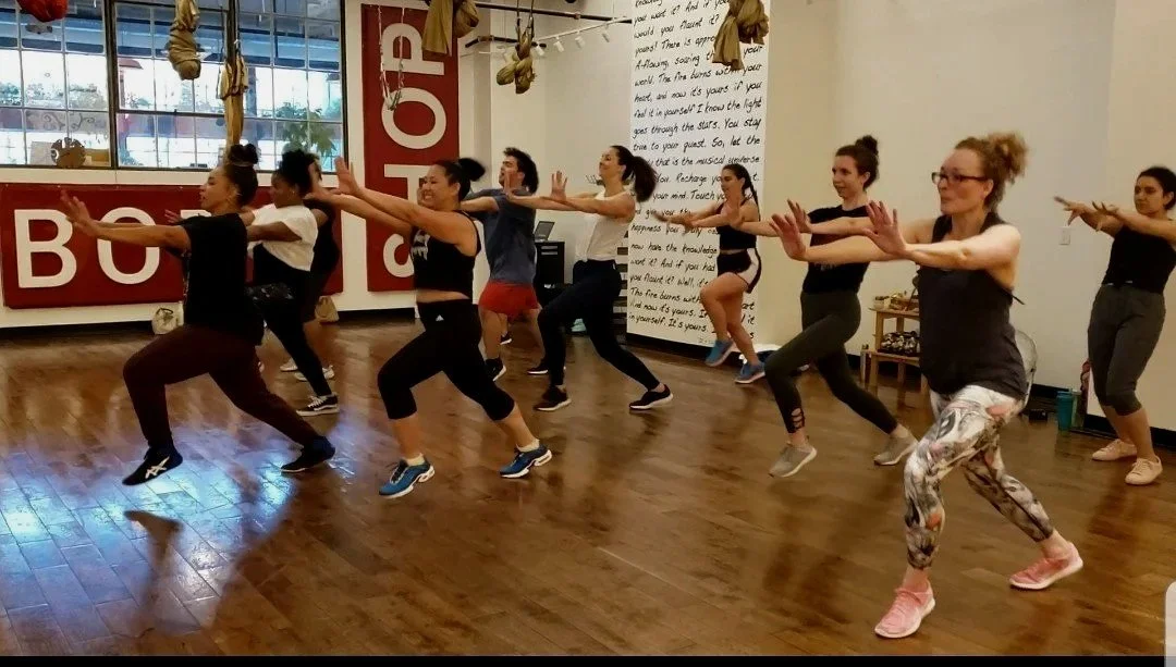 Group of women doing a dance or fitness class in a studio with large windows and hanging hammocks or aerial yoga equipment.