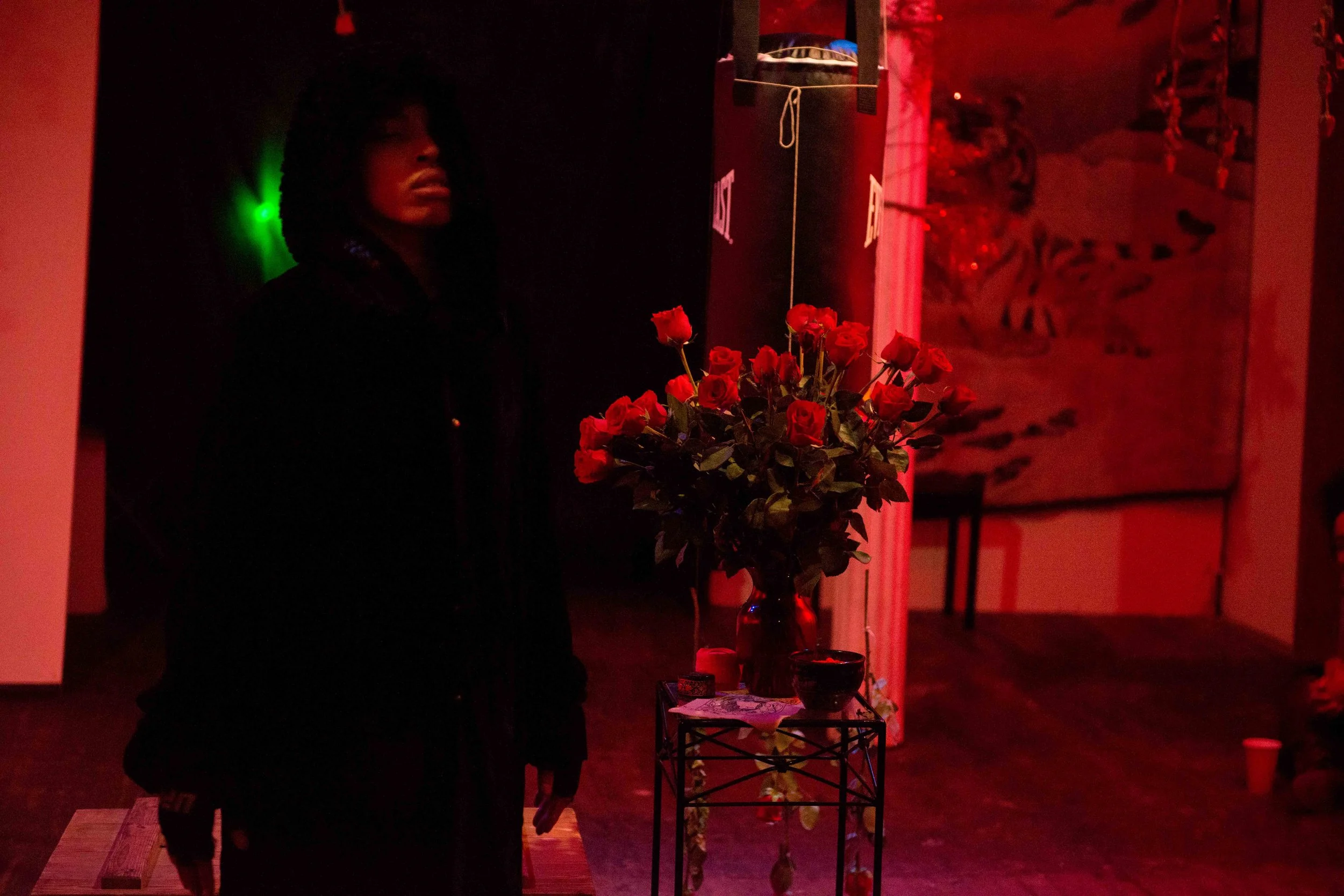 A woman stands in a dimly lit room with red lighting, near a table with a bouquet of red roses in a vase.