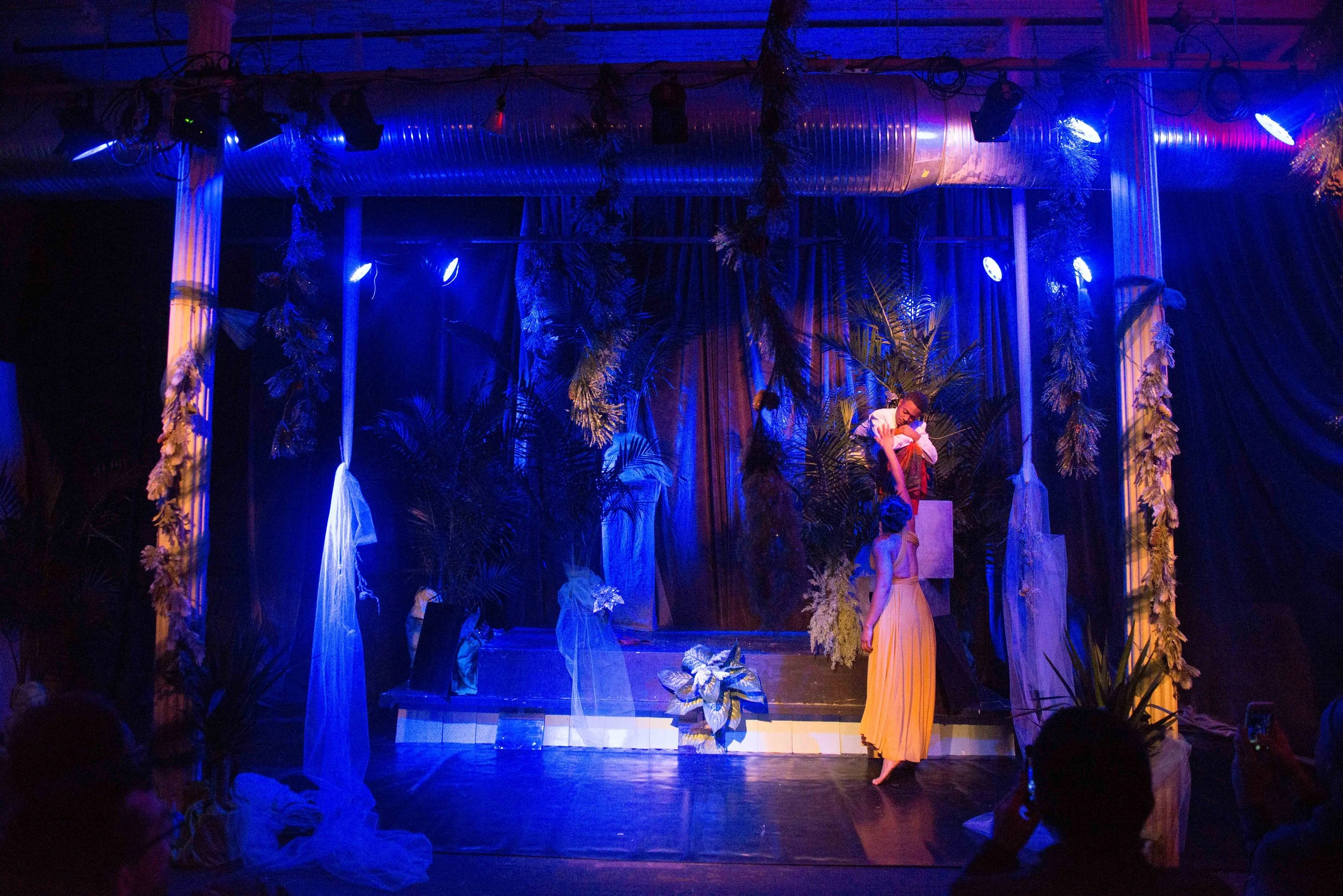 Theatrical stage with two actors, a woman and a man, surrounded by tropical plants, blue and purple lighting, with audience members taking photos.