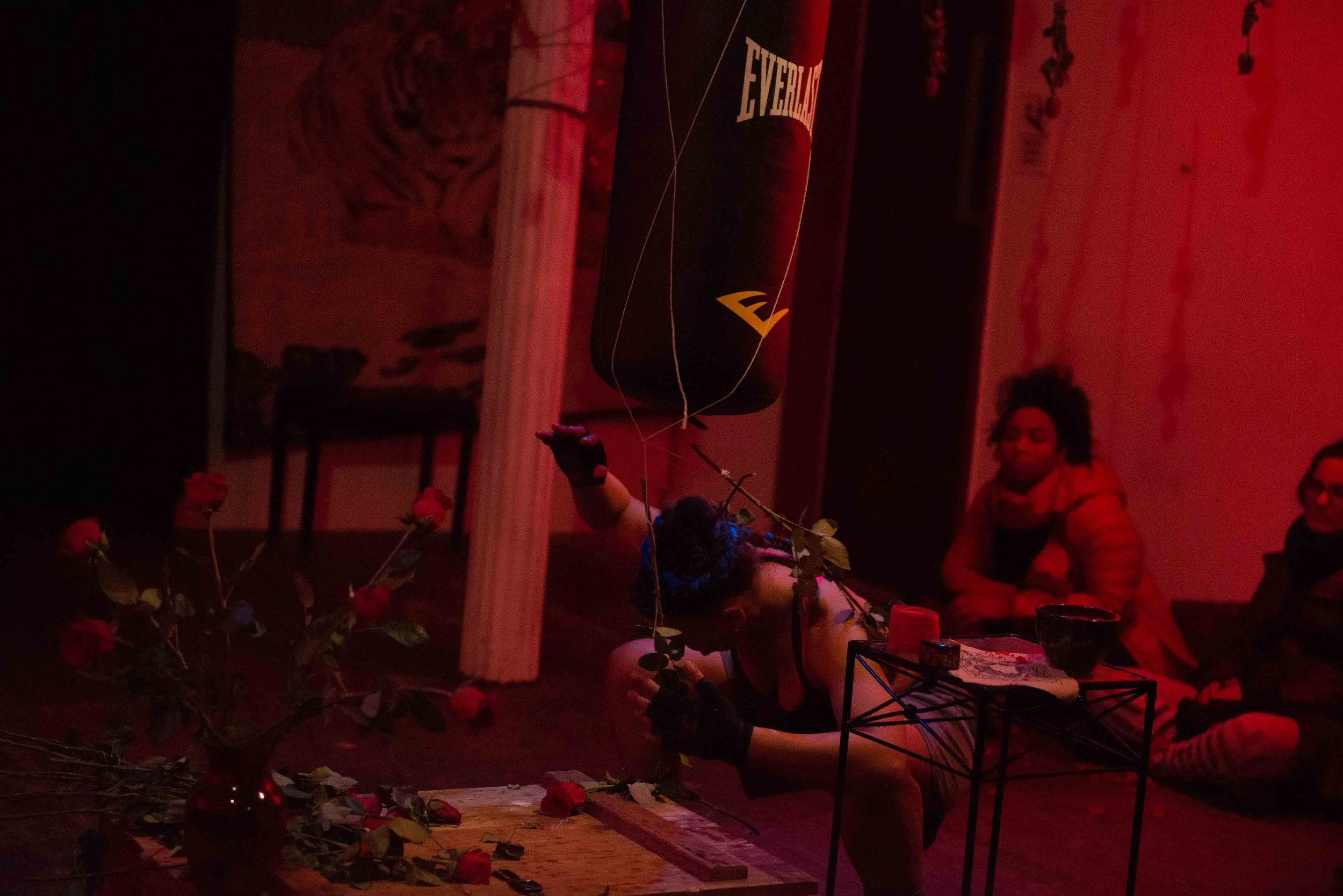 Performer doing a contortion act with roses, surrounded by an audience in a dimly lit room with red lighting.