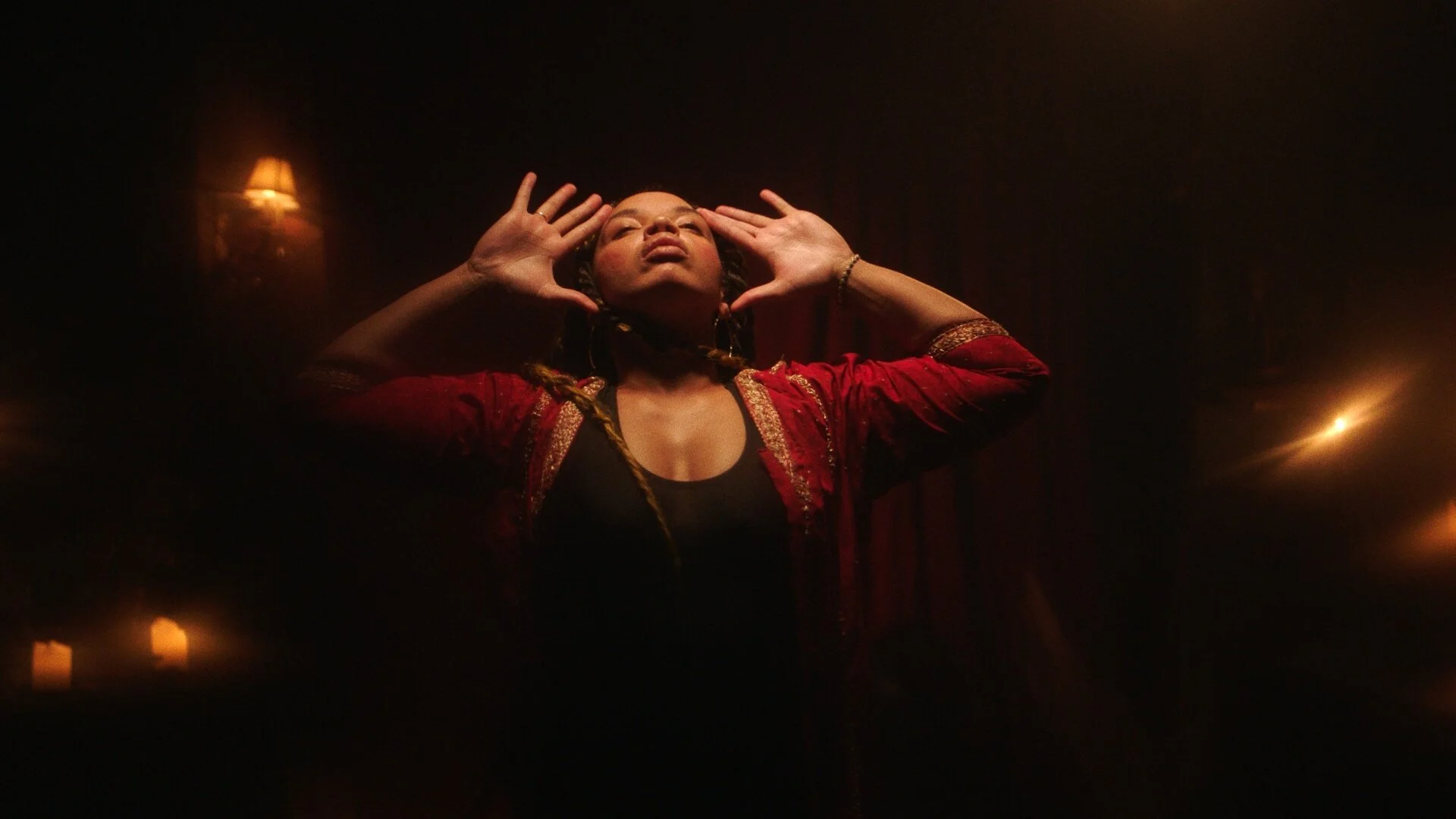 A woman dressed in a red and gold jacket with gold braid trims, standing against a dark background with warm lighting, holding her hands near her head with eyes closed in a dramatic or expressive pose.