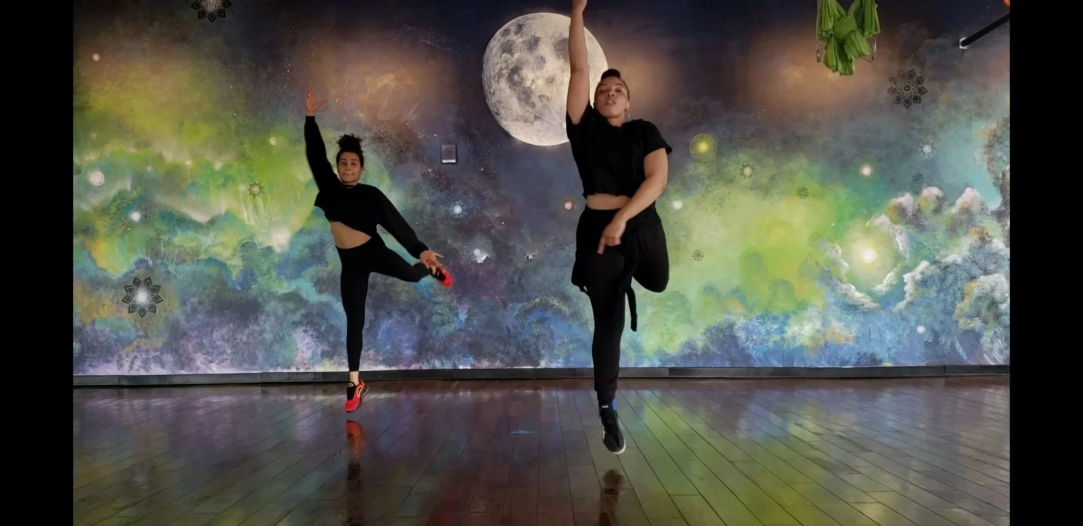 Two women dancing in front of a colorful space-themed mural that features the moon, stars, and nebulae. One woman is mid-air with her leg extended and arm raised, while the other is danced with her hand near her face, both wearing black outfits.
