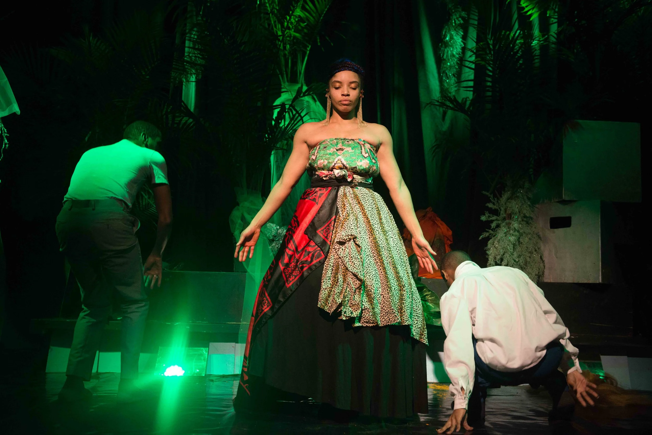 A woman standing on stage with her eyes closed, wearing a colorful strapless dress with a patterned fabric. Two men are kneeling on the stage near her, one on each side, and green stage lights illuminate the scene with a backdrop of green foliage.