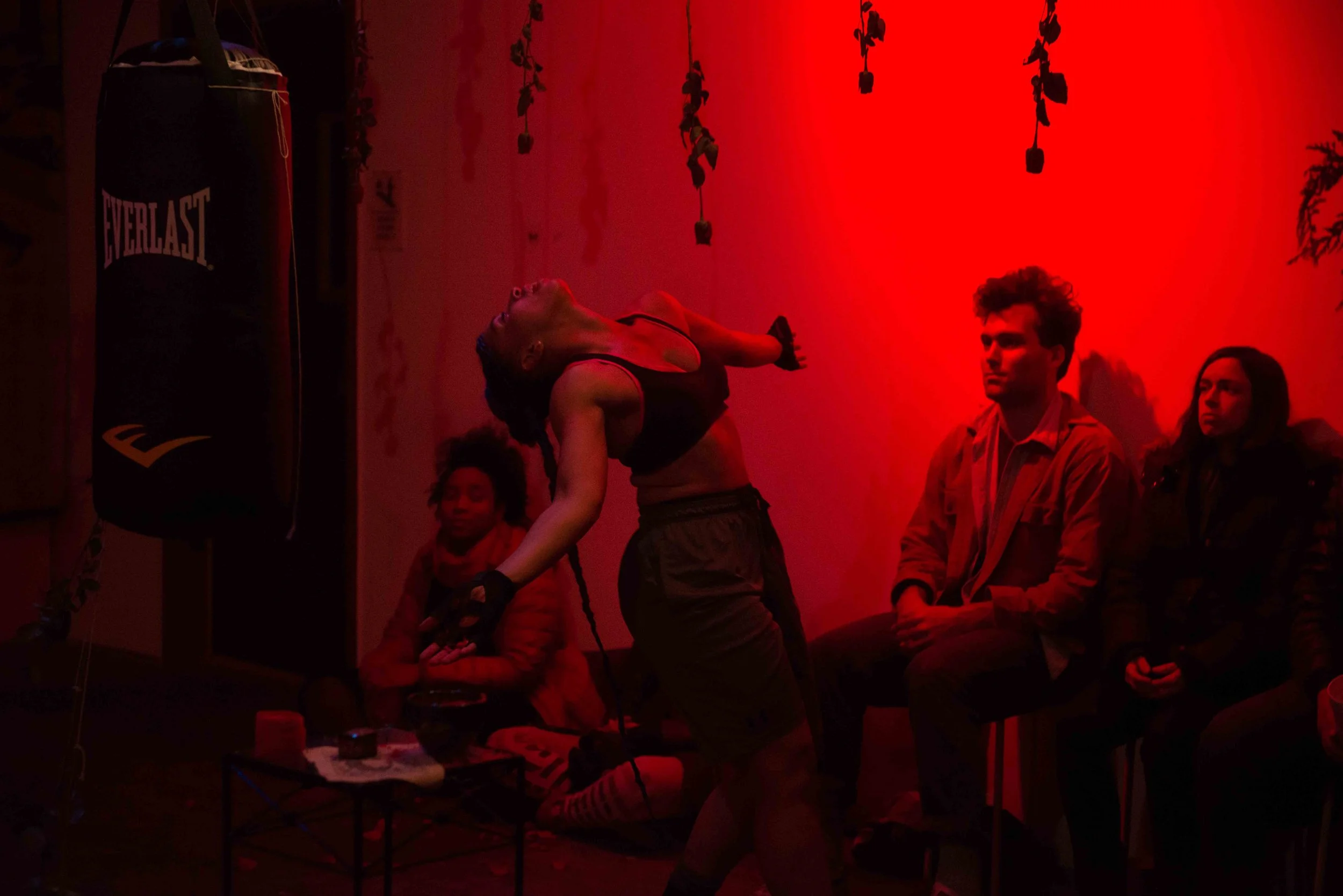 A woman in athletic wear, wearing gloves, is practicing punching a black punching bag in a fitness studio with red lighting. Three people are seated on a bench observing her, two women and one man, with a red wall and hanging plants in the background.