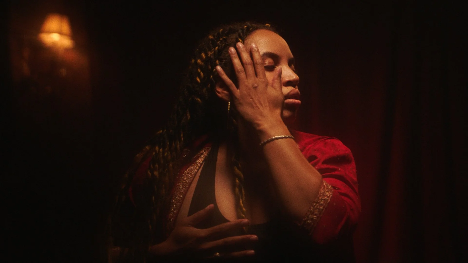 A woman with braided hair and wearing a red garment is performing a dance move in a dimly lit room with warm lighting.