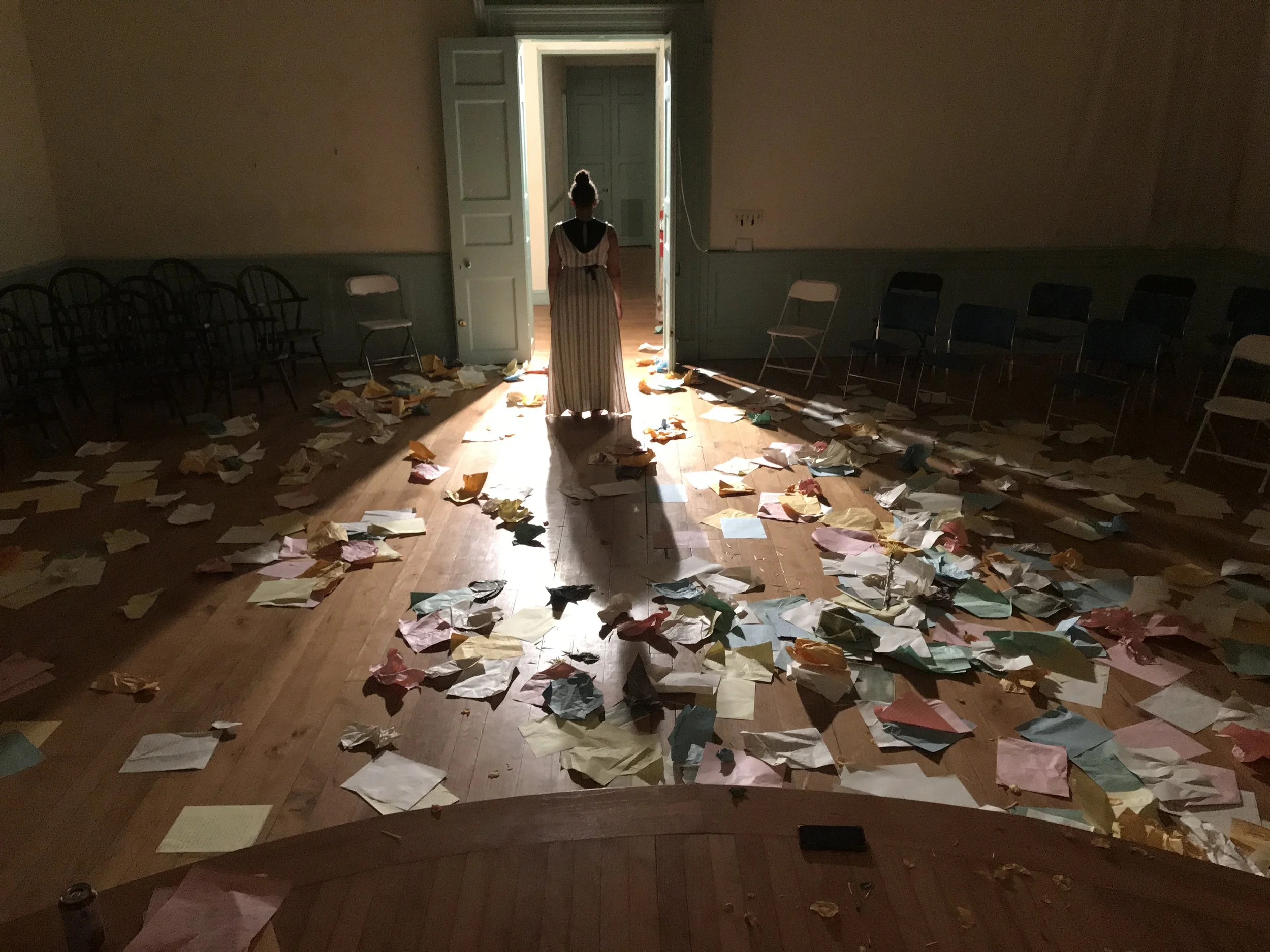 A room with scattered papers on the wooden floor, a woman in period clothing standing in the doorway, and unused chairs lining the walls.