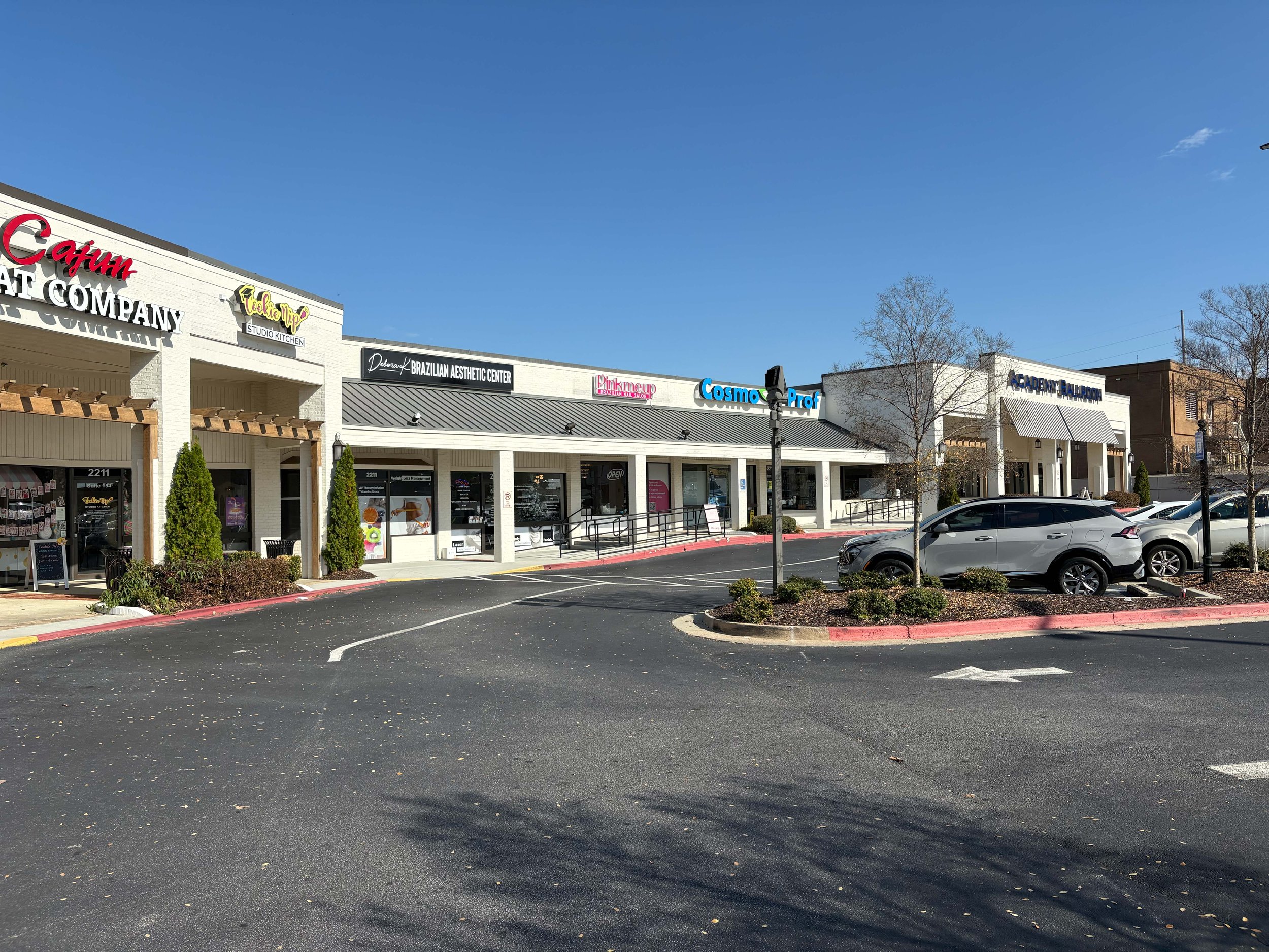 A shopping plaza with several stores, including Cafun, Studio, Brazilian Aesthetic Center, Pinkmup, Cosmo Prof, and Academy Ballroom. There are parked cars, a small landscaped area with trees and bushes, and a parking lot with clear parking spaces, u