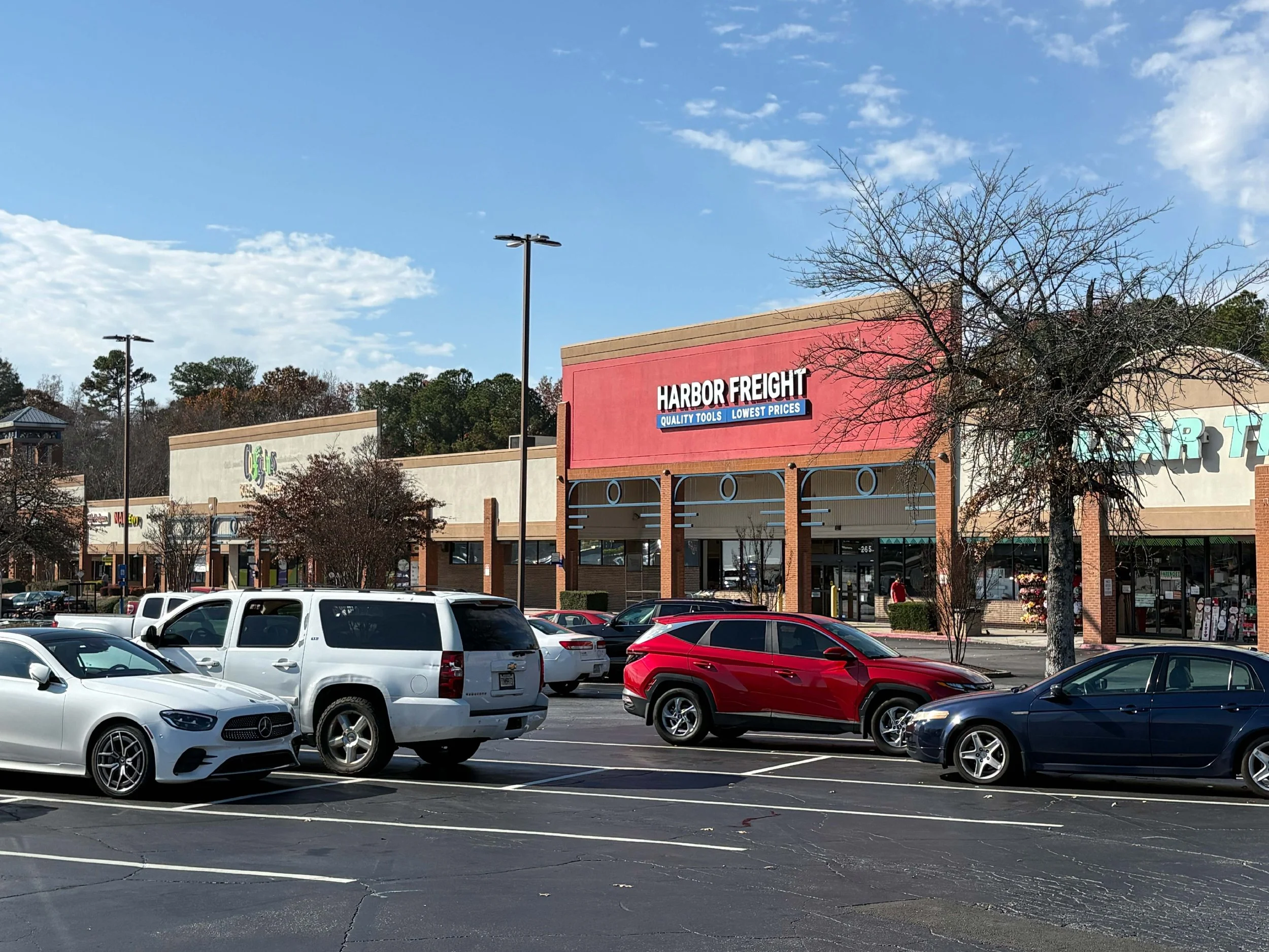 A parking lot in front of a shopping plaza with cars parked, including white, red, blue, and black vehicles. The background shows storefronts with signs, including 'Harbor Freight' in a red building, under a partly cloudy blue sky.